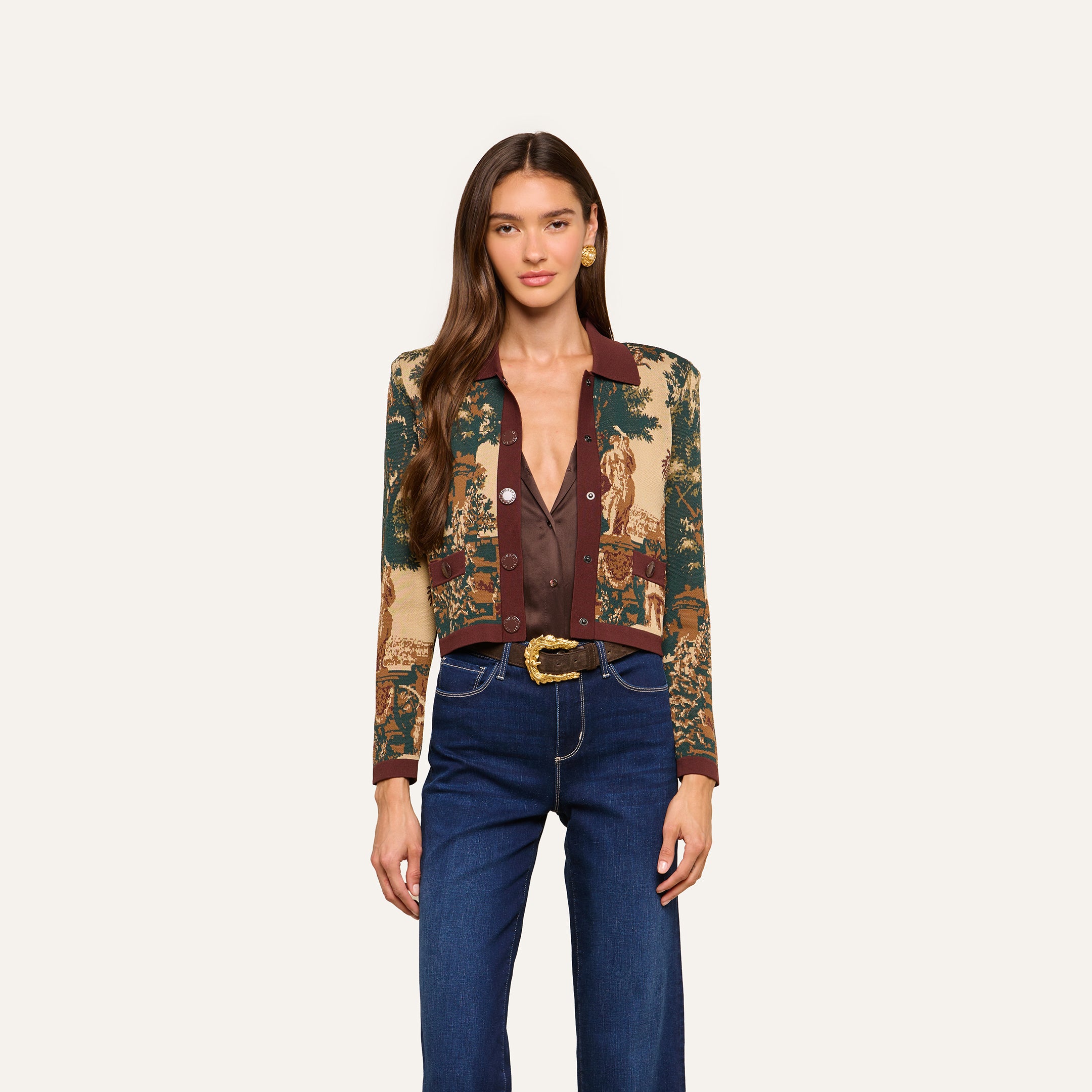 A woman with long brown hair wears the Osiris Jacquard Cardigan featuring a tapestry-inspired print with nature scenes, styled with a brown blouse, blue high-waisted jeans, and a belt with a gold buckle, standing against a plain off-white background.