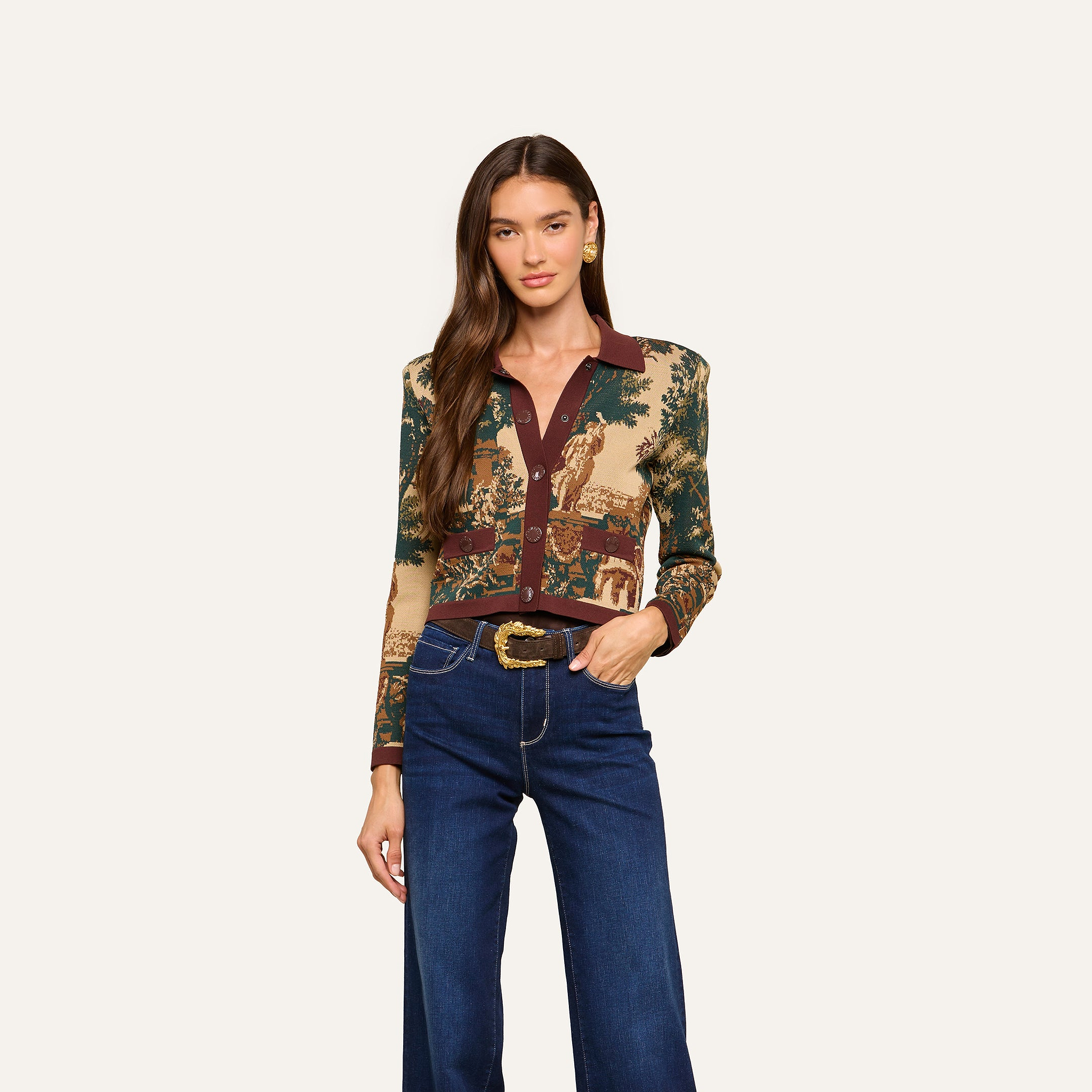 A woman with long brown hair stands against a plain white background, looking at the camera with a relaxed expression. She is wearing the Osiris Jacquard Cardigan, which features a patterned jacquard knit and a brown collar, paired with wide-leg blue jeans.