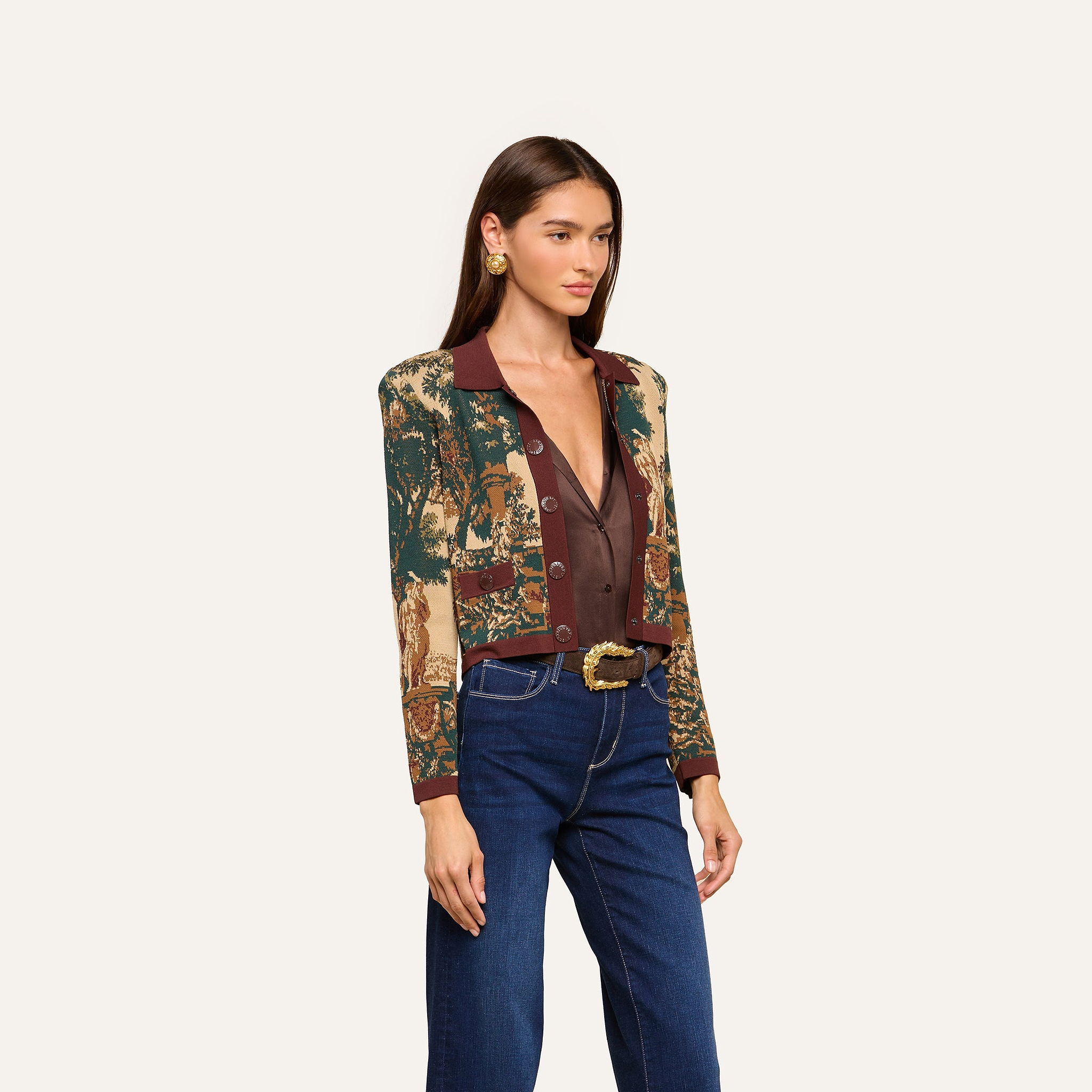 A woman with straight brown hair wears the Osiris Jacquard Cardigan, styled over a deep V-neck brown top, blue jeans, and a belt with a decorative buckle, standing against a plain light background.