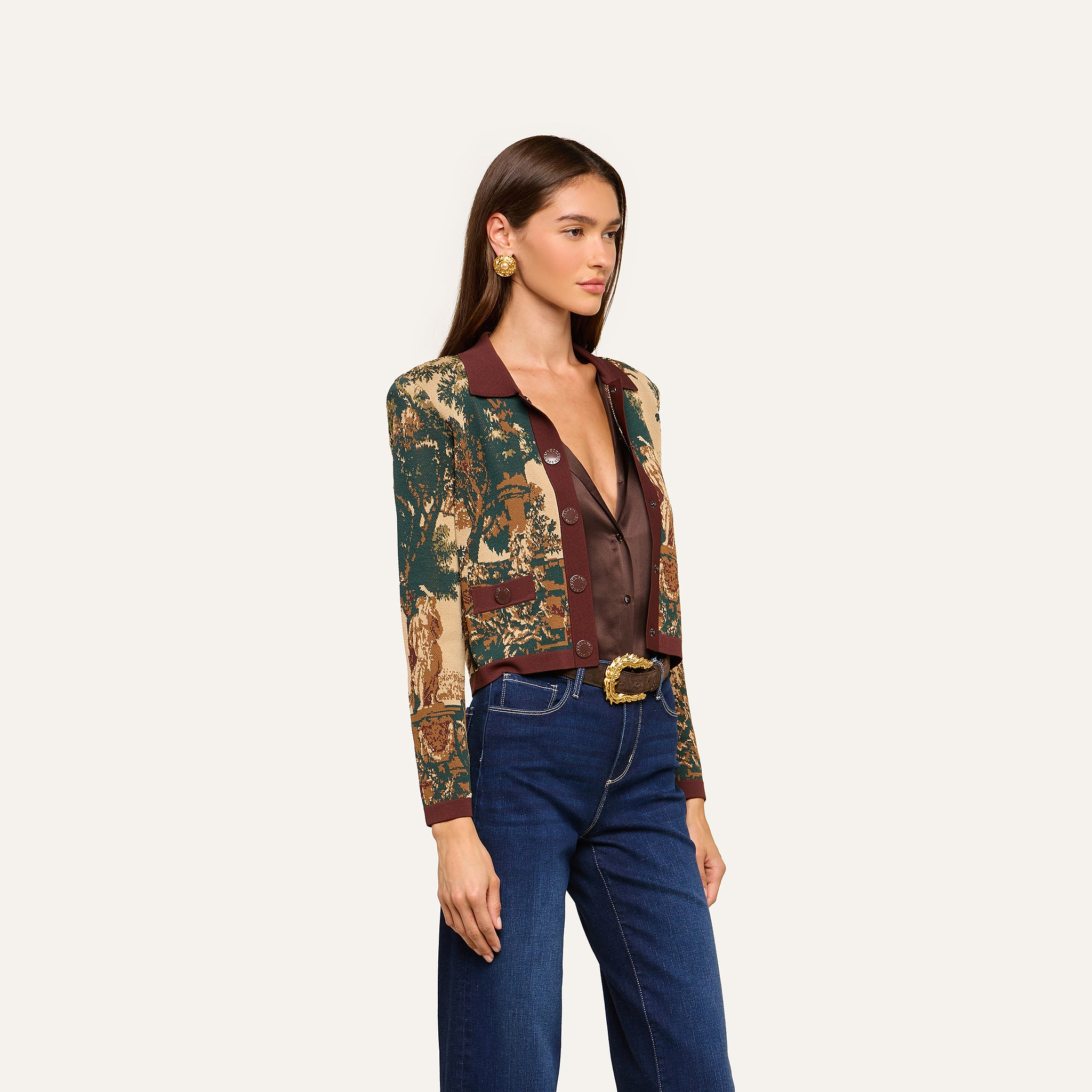 Wearing the Osiris Jacquard Cardigan, a woman with long brown hair pairs it with a sheer brown blouse, blue jeans, gold earrings, and a decorative belt as she stands against a plain light background.