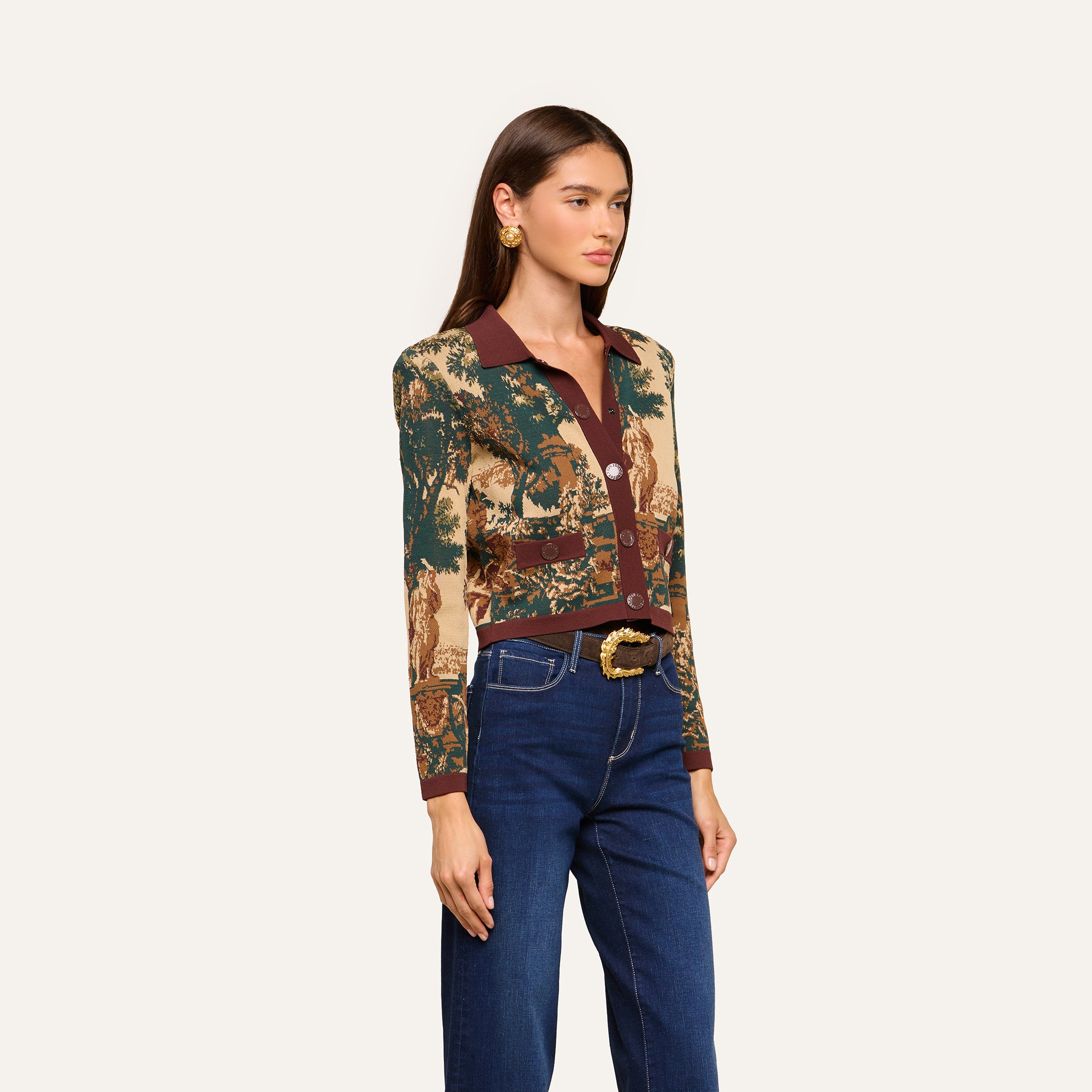 A woman with straight brown hair wears the Osiris Jacquard Cardigan, blue jeans, a black belt with a large gold buckle, and gold earrings, standing against a plain light background.