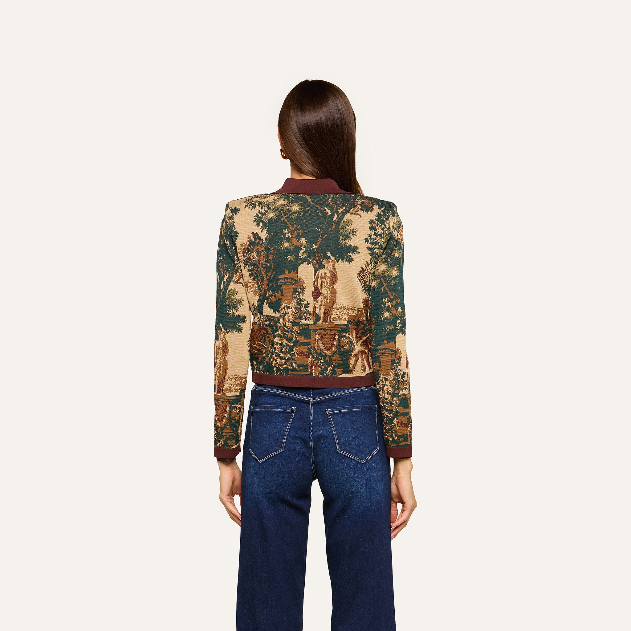 A woman with straight brown hair, seen from behind, wears the Osiris Jacquard Cardigan with a nature scene motif and blue jeans, standing against a plain light background.