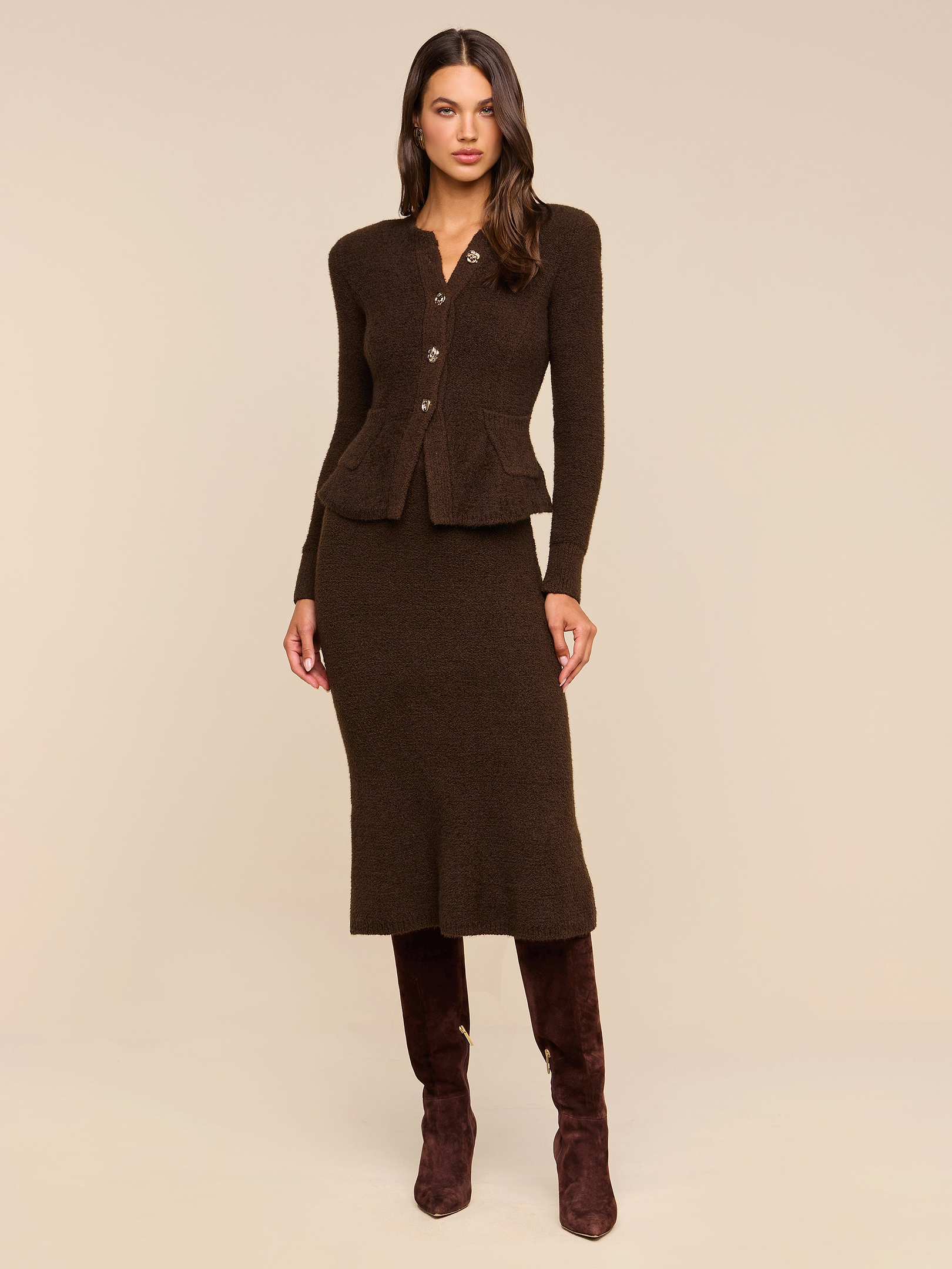 Woman stands against a plain white background wearing a dark brown buttoned cardigan, the Salem Knit Skirt, and brown knee-high boots.