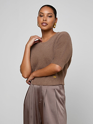 A woman with medium skin tone stands against a plain background, wearing the Guinevere Cropped Pullover—an ultra soft knit brown crewneck sweater—paired with tan satin trousers. She accessorizes with gold hoop earrings and has her hair pulled back, her right arm bent with her hand near her neck.