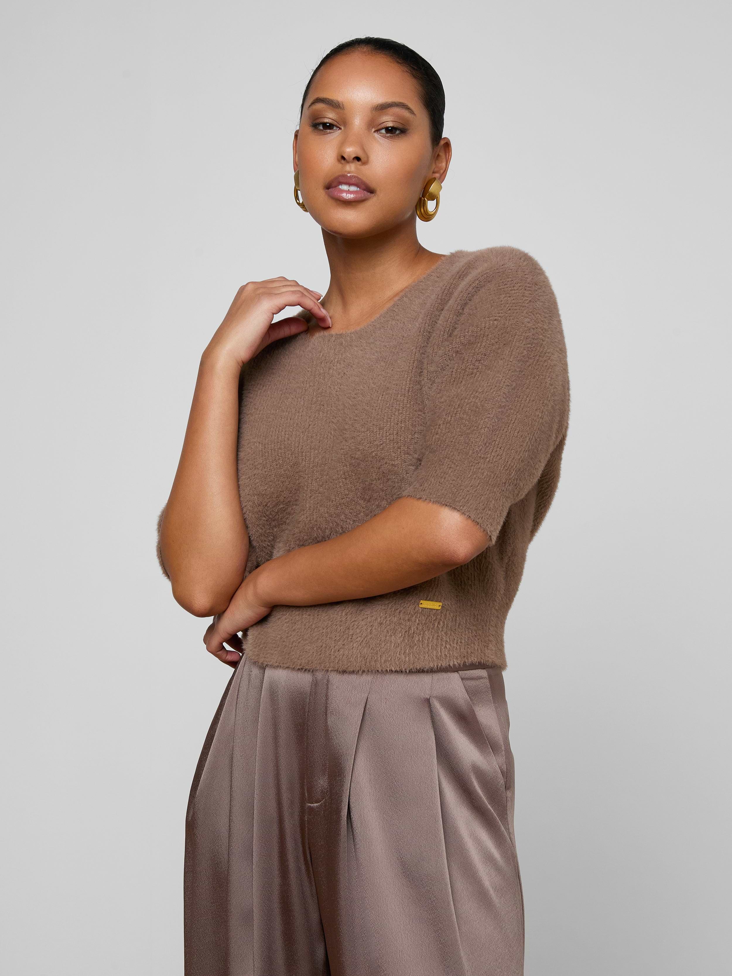 A woman with medium skin tone stands against a plain background, wearing the Guinevere Cropped Pullover—an ultra soft knit brown crewneck sweater—paired with tan satin trousers. She accessorizes with gold hoop earrings and has her hair pulled back, her right arm bent with her hand near her neck.