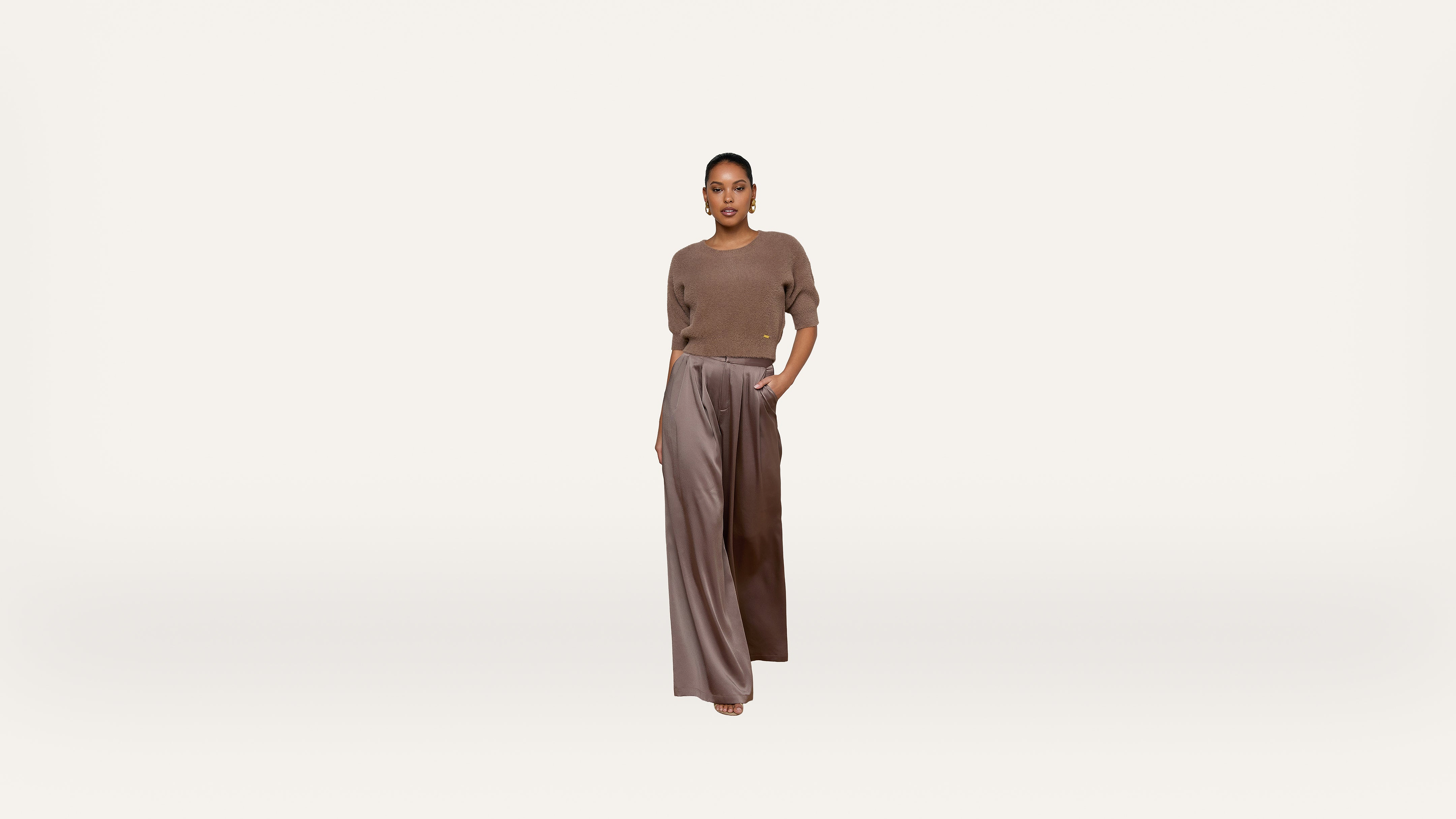 A woman stands against a plain white background, wearing a brown short-sleeve sweater and wide-legged, high-waisted taupe pants. She has one hand in her pocket and looks confidently at the camera.