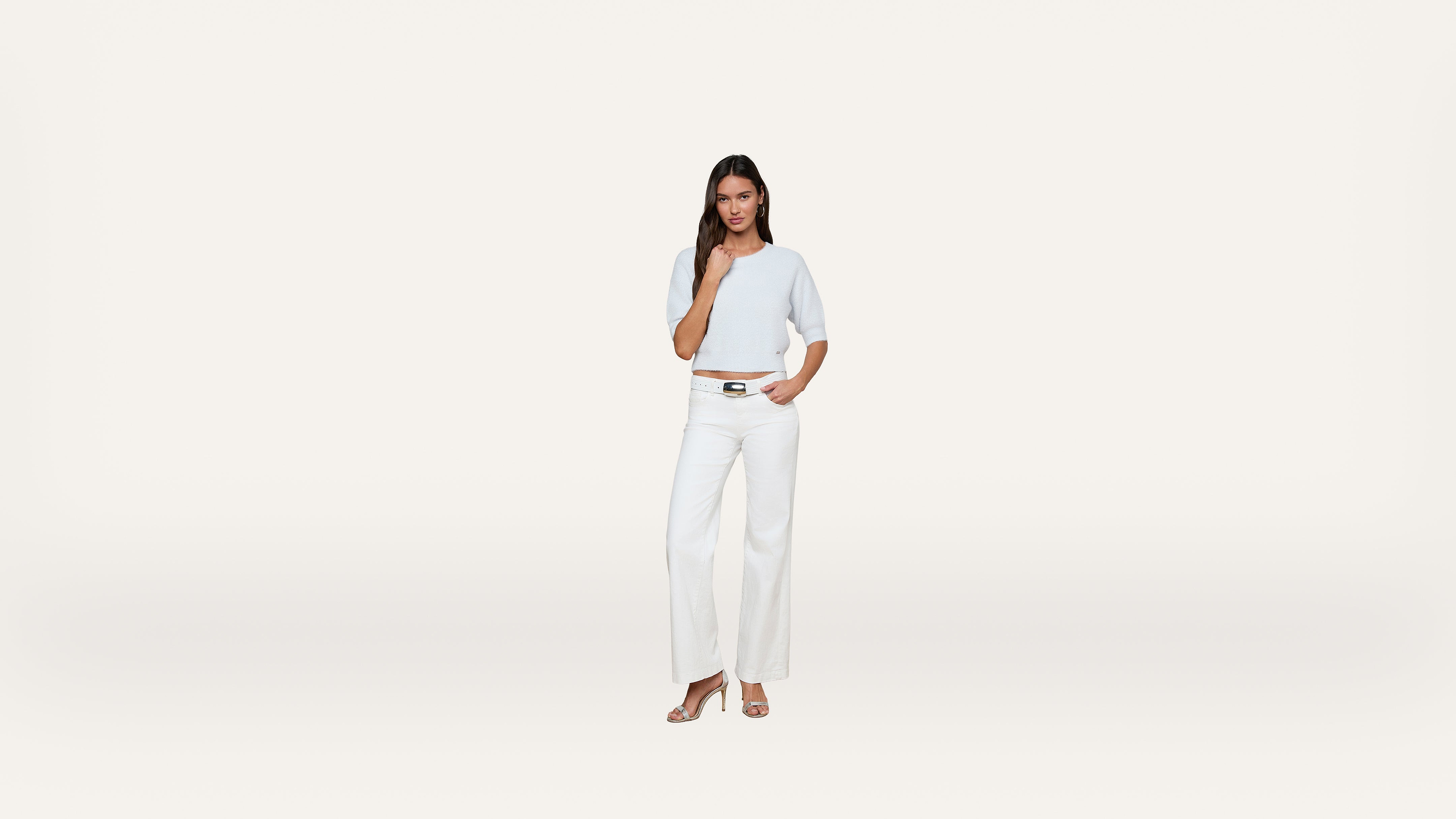 A woman in a light blue top and white wide-leg pants stands against a plain white background, with one hand touching her hair and the other resting by her side.