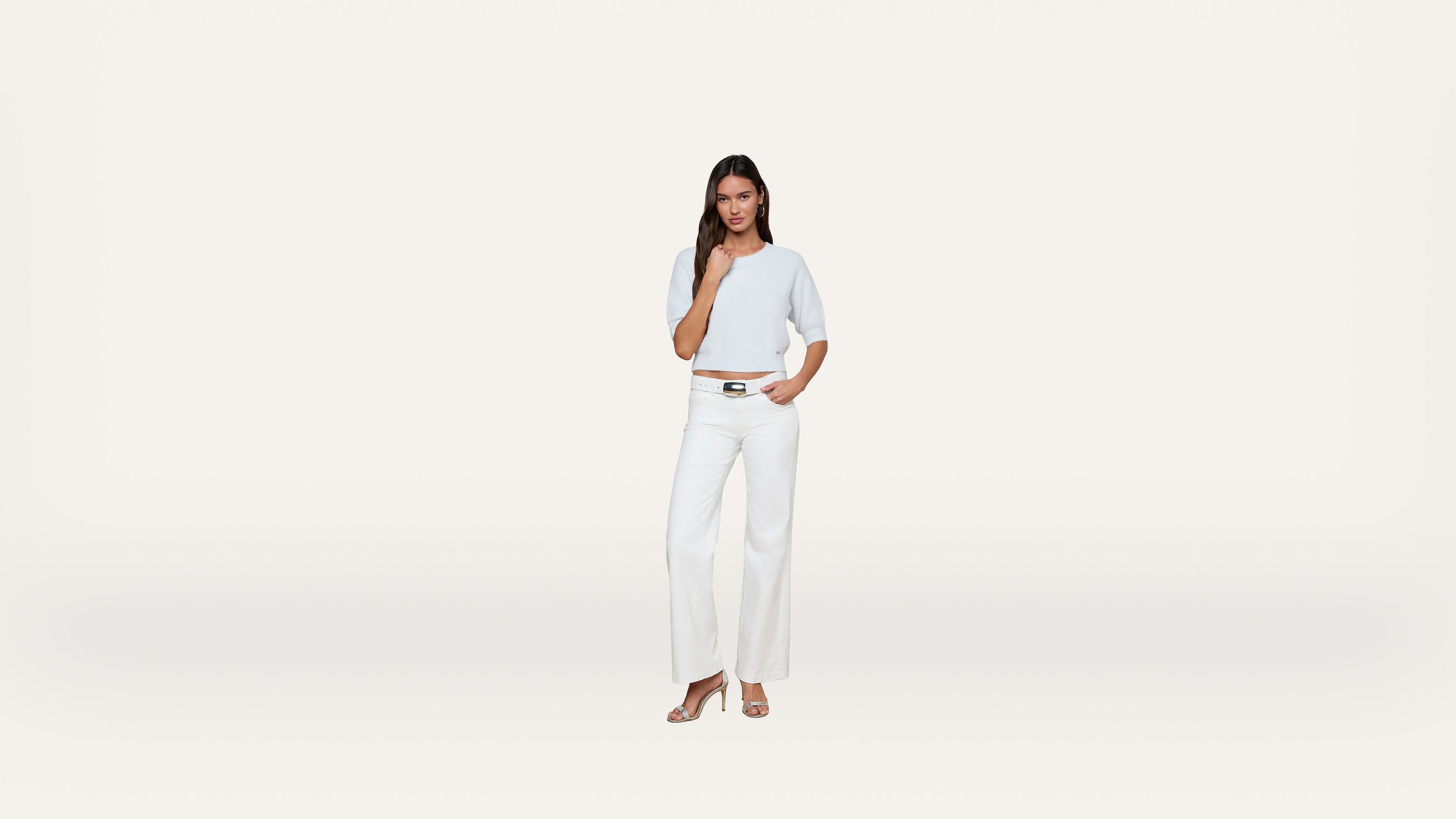 A woman in a light blue top and white wide-leg pants stands against a plain white background, with one hand touching her hair and the other resting by her side.