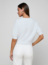 A woman stands with her back to the camera, wearing the Guinevere Cropped Pullover—a white soft knit crewneck sweater—paired with white jeans. She has straight brown hair, hoop earrings, and one hand resting on her hip against a plain, light-colored background.