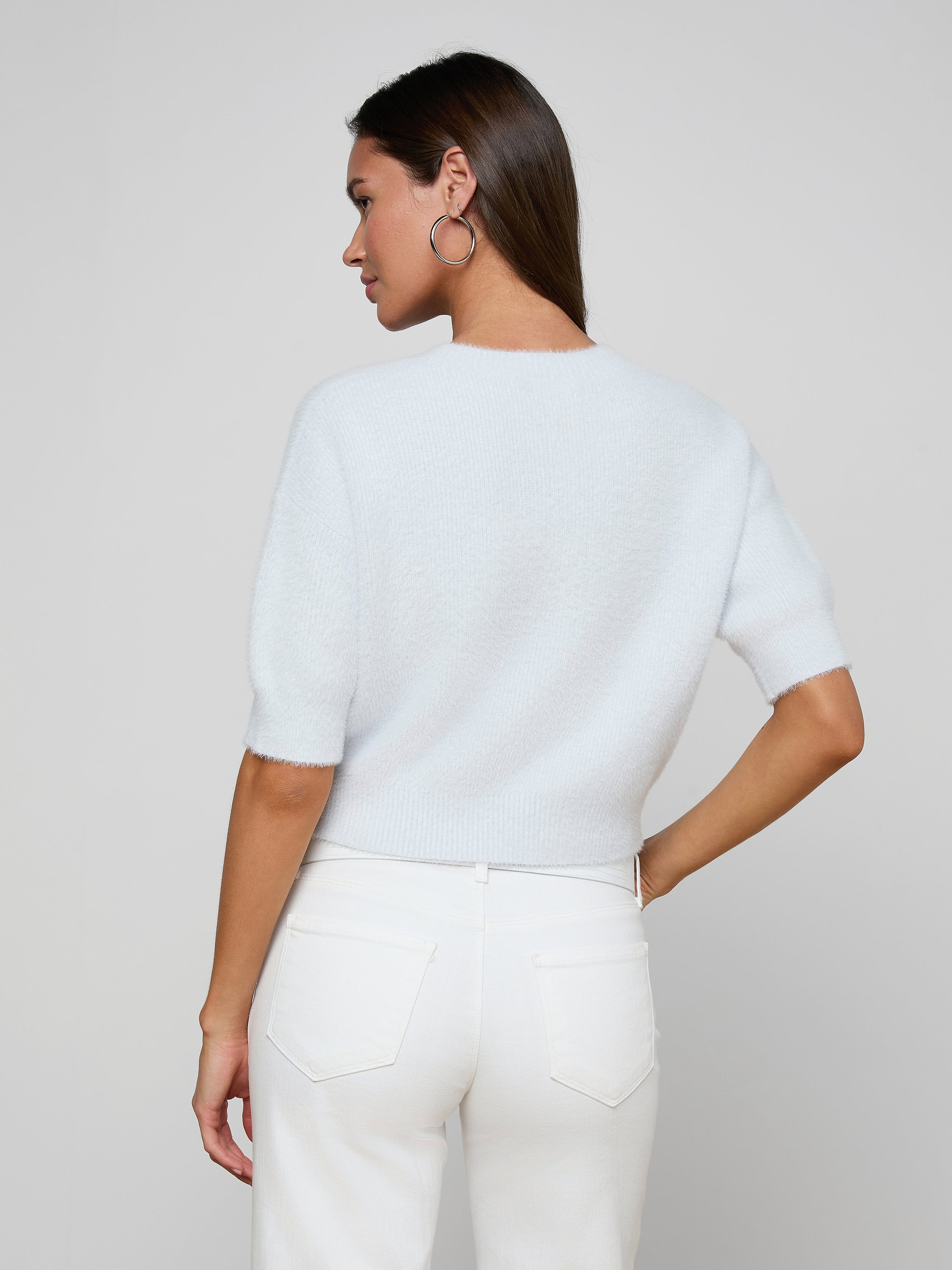 A woman stands with her back to the camera, wearing the Guinevere Cropped Pullover—a white soft knit crewneck sweater—paired with white jeans. She has straight brown hair, hoop earrings, and one hand resting on her hip against a plain, light-colored background.