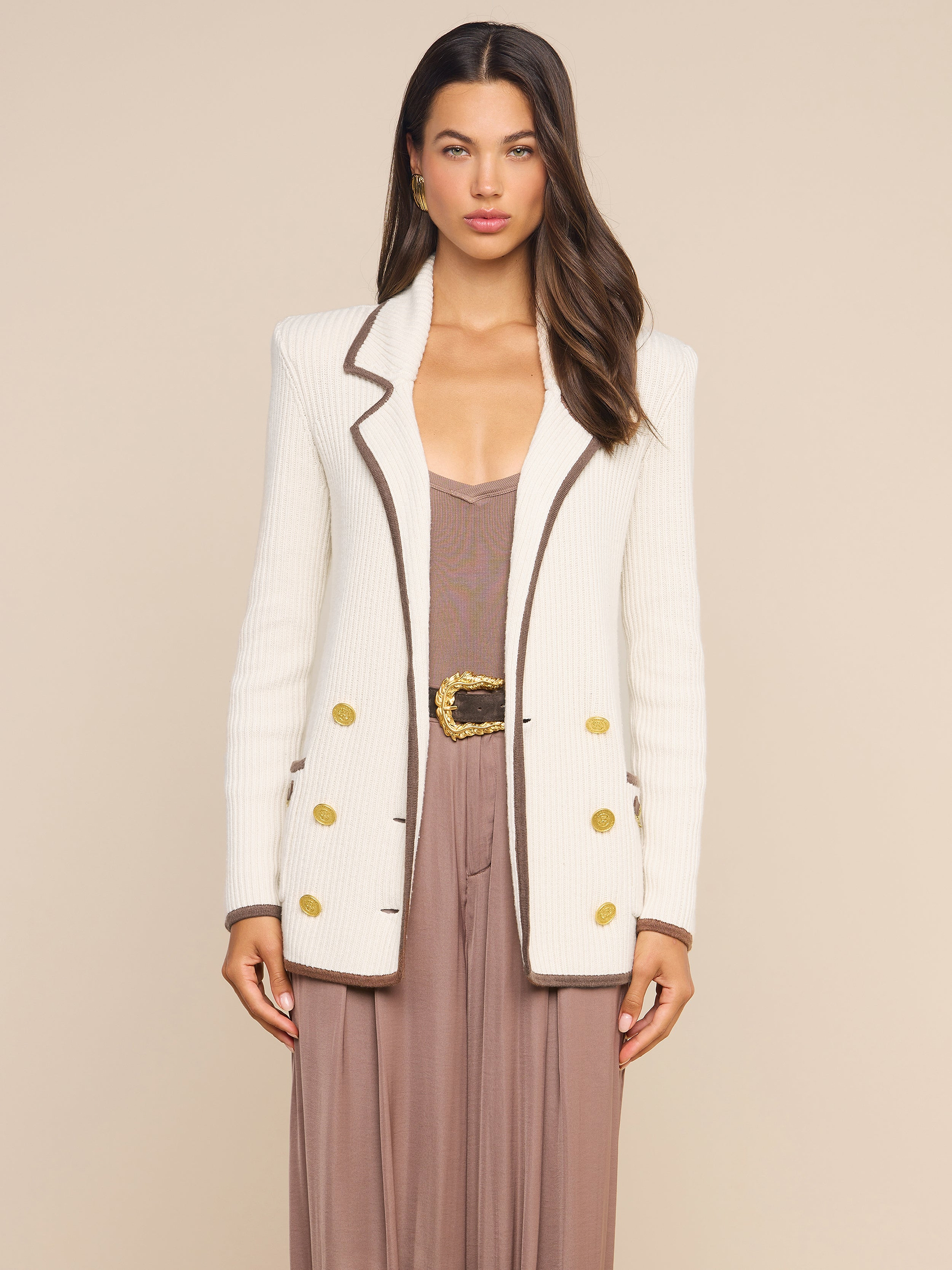 A woman stands facing forward, wearing the Ranch Knit Blazer—a cream, double-breasted style with gold buttons—over a taupe dress and belt. She has long, wavy brown hair and a neutral expression against a plain background.