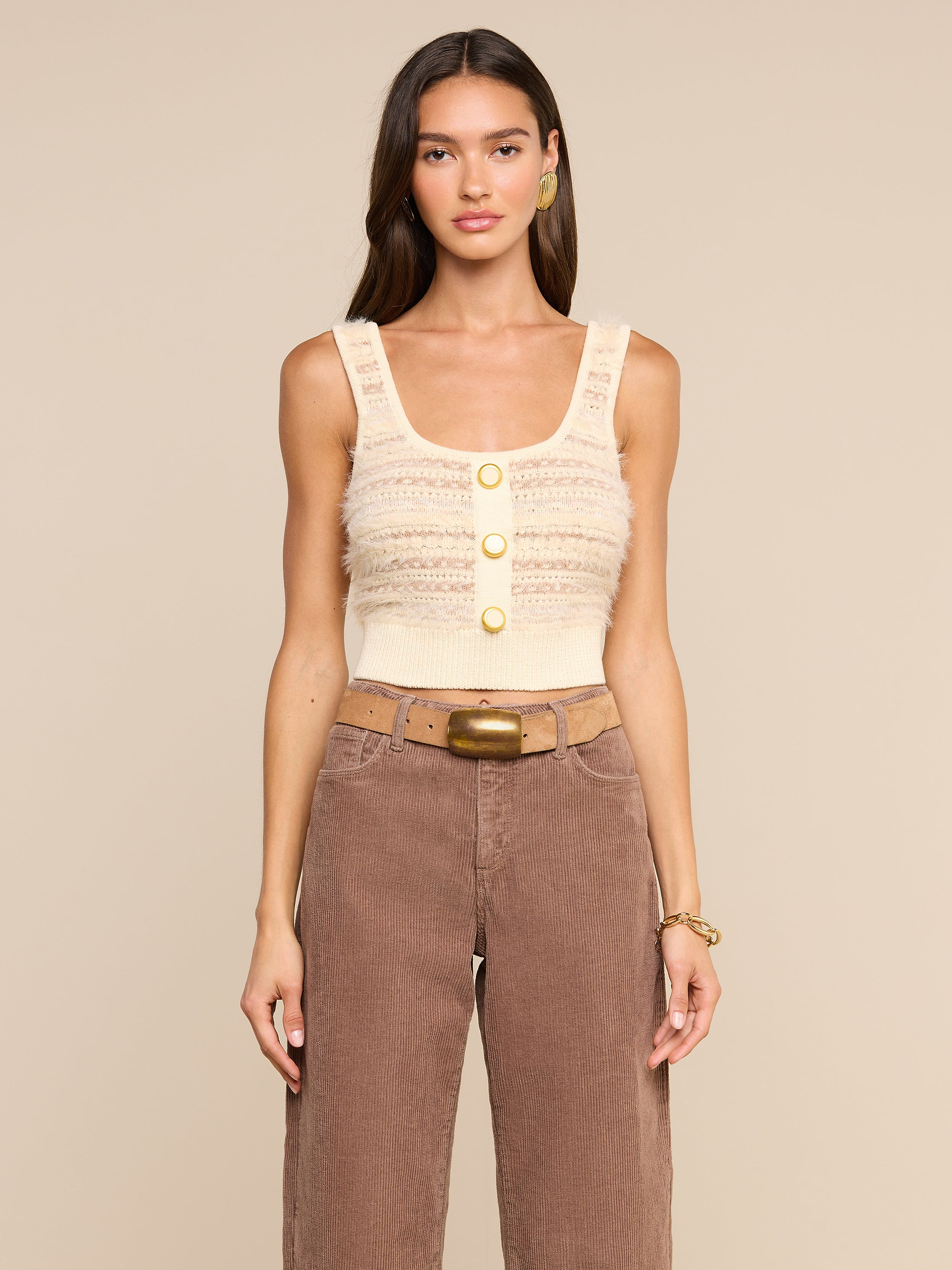 A woman wears the Galene Tank, a sleeveless beige cropped knit top with three decorative buttons, paired with brown high-waisted pants, a wide belt with a brass buckle, gold earrings, and a bracelet, as she stands against an off-white background.