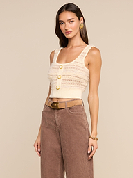 A woman wearing the Galene Tank—a cropped knit top with eyelash texture, three decorative buttons, and wide straps—paired with high-waisted brown corduroy pants and a tan belt, poses against a plain background.