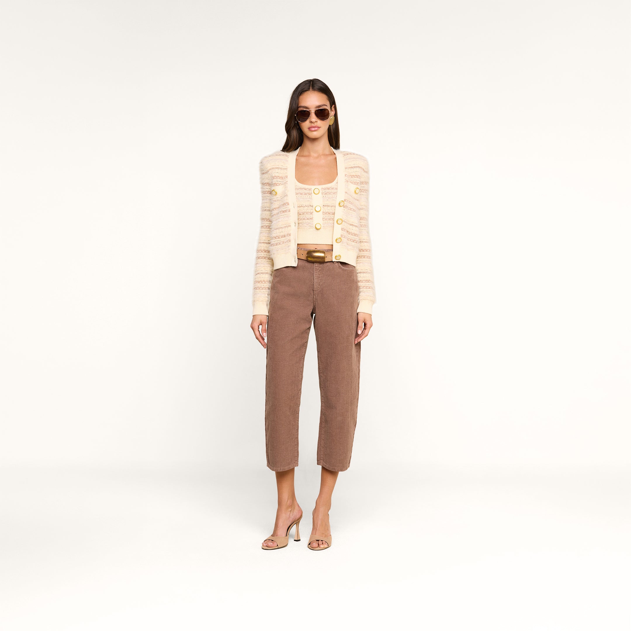 A woman wearing sunglasses, a beige striped cardigan with decorative buttons over the Galene Tank, brown cropped pants, and beige high-heeled sandals stands against a plain white background.