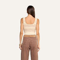 A woman with long brown hair is seen from behind, wearing the cream cropped Galene Tank featuring decorative buttons and high-waisted brown pants, standing against a plain light background.