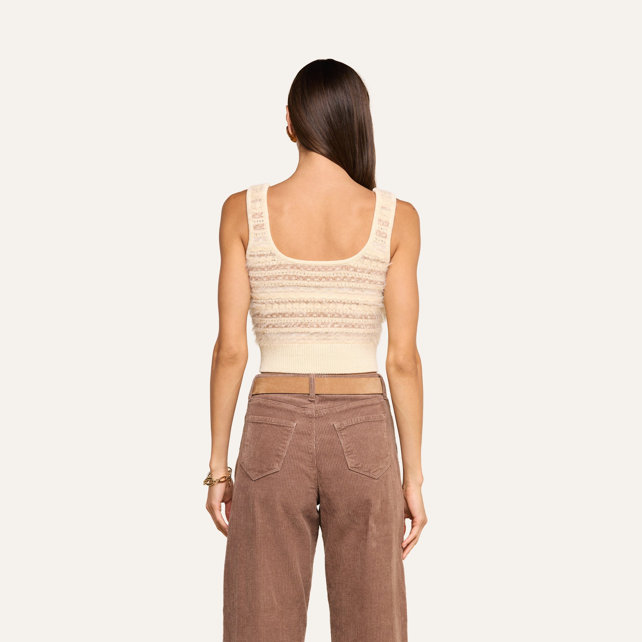 A woman with long brown hair is seen from behind, wearing the cream cropped Galene Tank featuring decorative buttons and high-waisted brown pants, standing against a plain light background.