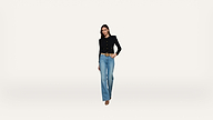 A woman stands against a plain white background wearing a black blazer, blue jeans, and brown shoes, with one hand in her pocket and a slight smile.