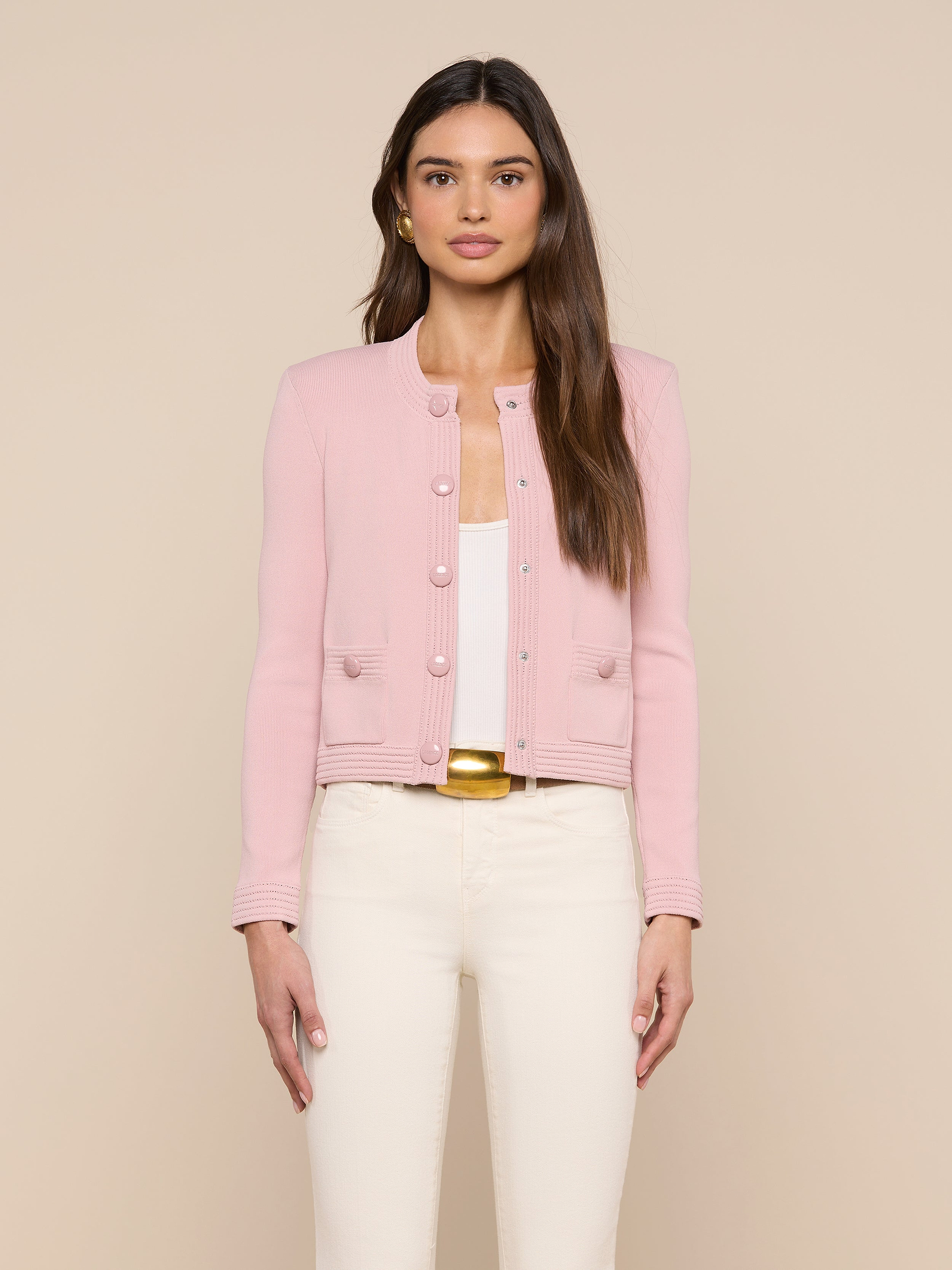 A woman stands facing forward, wearing the Patsy Cropped Cardigan in light pink pointelle knit over a white top and cream-colored pants, with long brown hair against a neutral background.