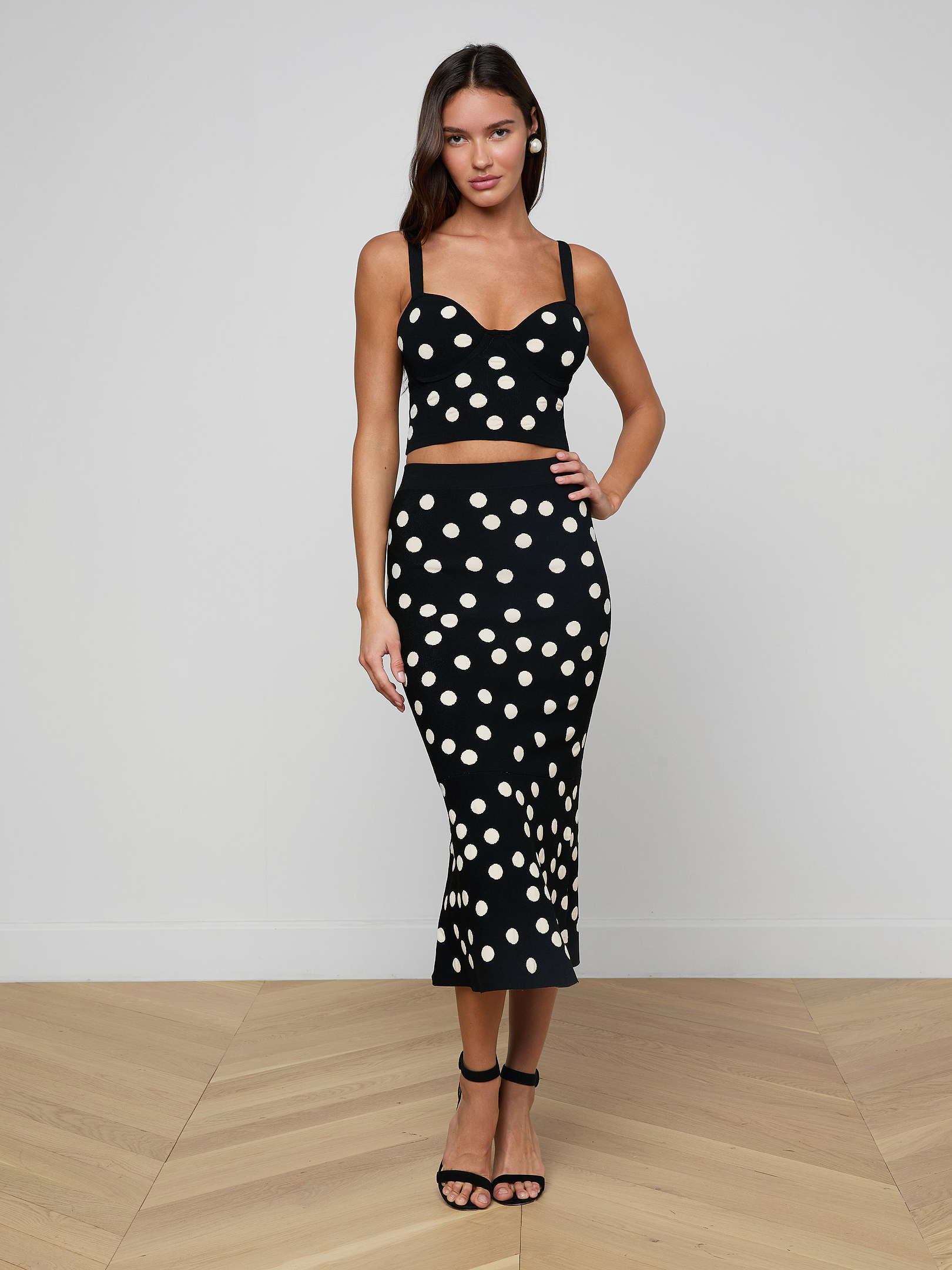 A woman stands against a plain, light background wearing a black, sleeveless crop top paired with the Mae Knit Flare Skirt—a high-waisted, polka dot fit and flare skirt—and black sandals.