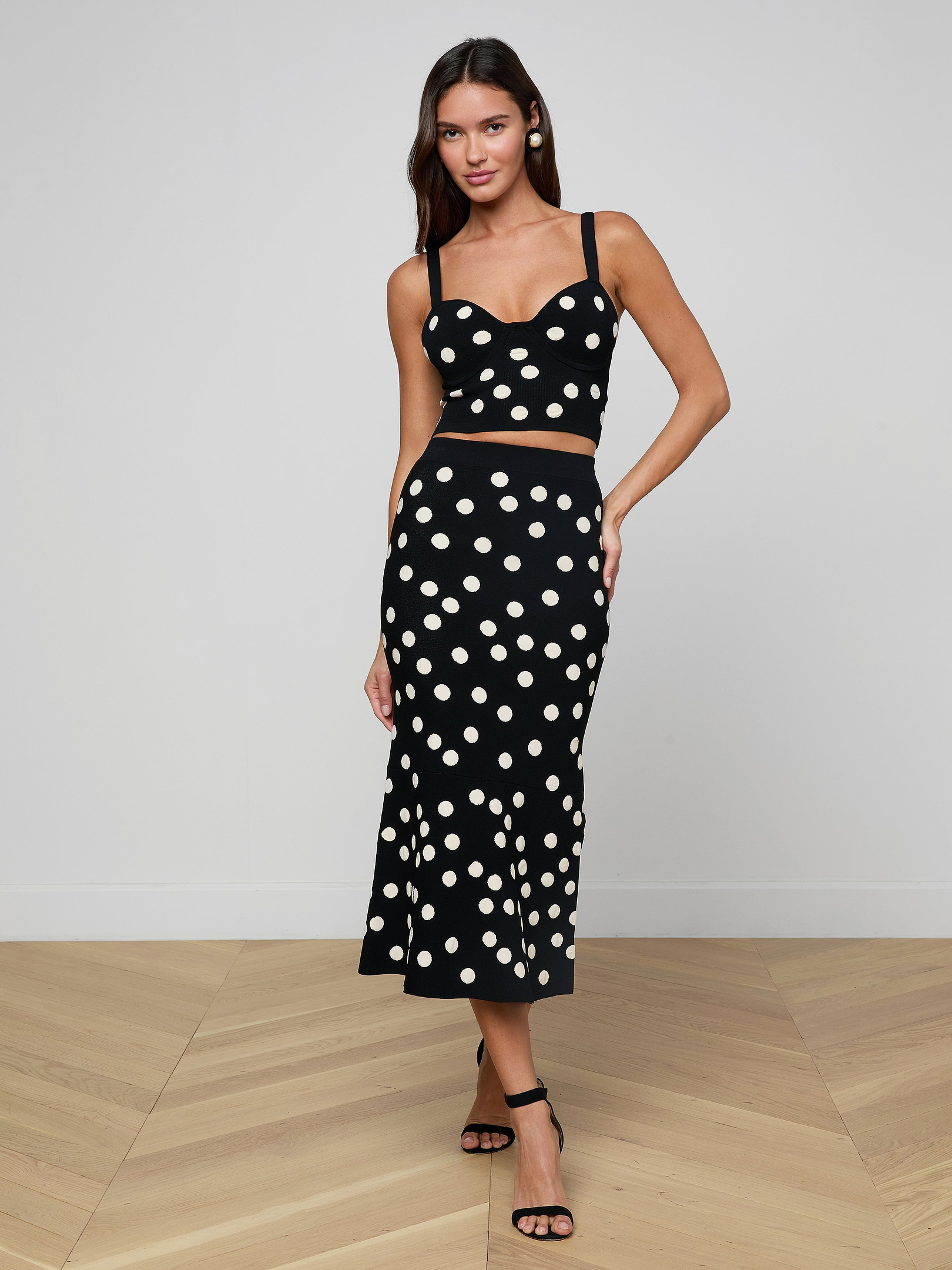 A woman poses against a plain white background wearing the Mae Knit Flare Skirt, a black skirt with large white polka dots, paired with a matching crop top and black strappy heels.