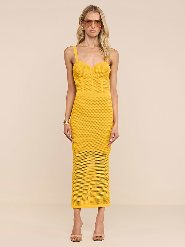 A woman wearing the Michelle Knit Dress—a sleeveless, fitted yellow midi dress featuring a sheer skirt and built-in bra—stands against a plain white background. She has long blonde hair, sunglasses, and is wearing sandals.
