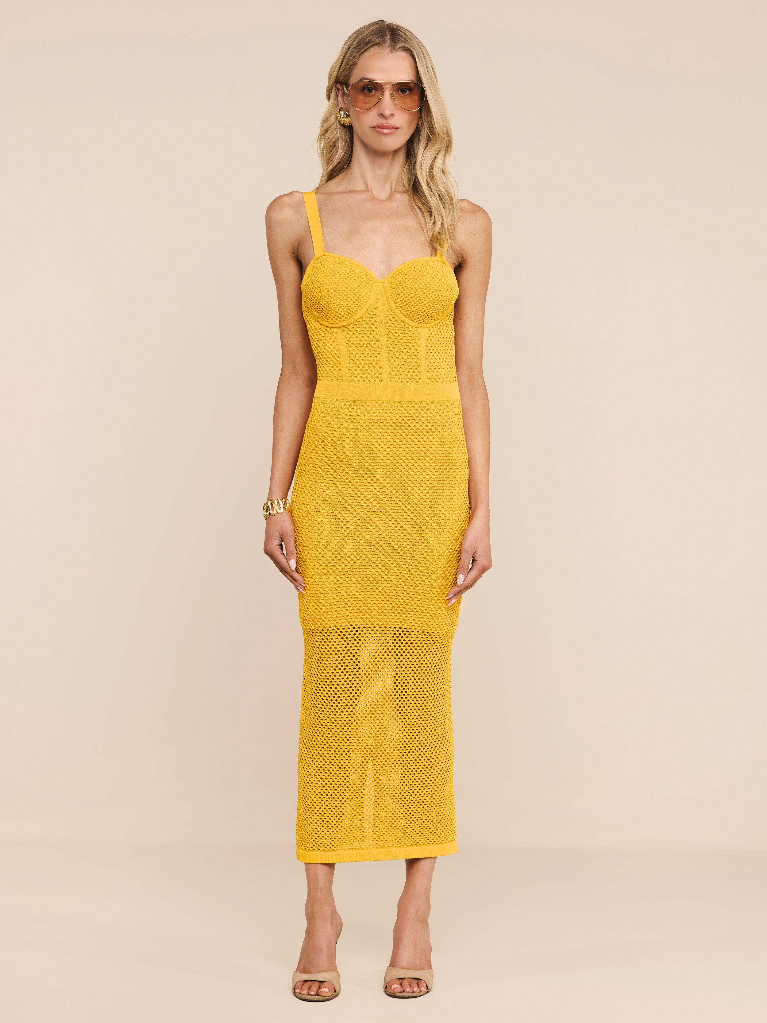A woman wearing the Michelle Knit Dress—a sleeveless, fitted yellow midi dress featuring a sheer skirt and built-in bra—stands against a plain white background. She has long blonde hair, sunglasses, and is wearing sandals.