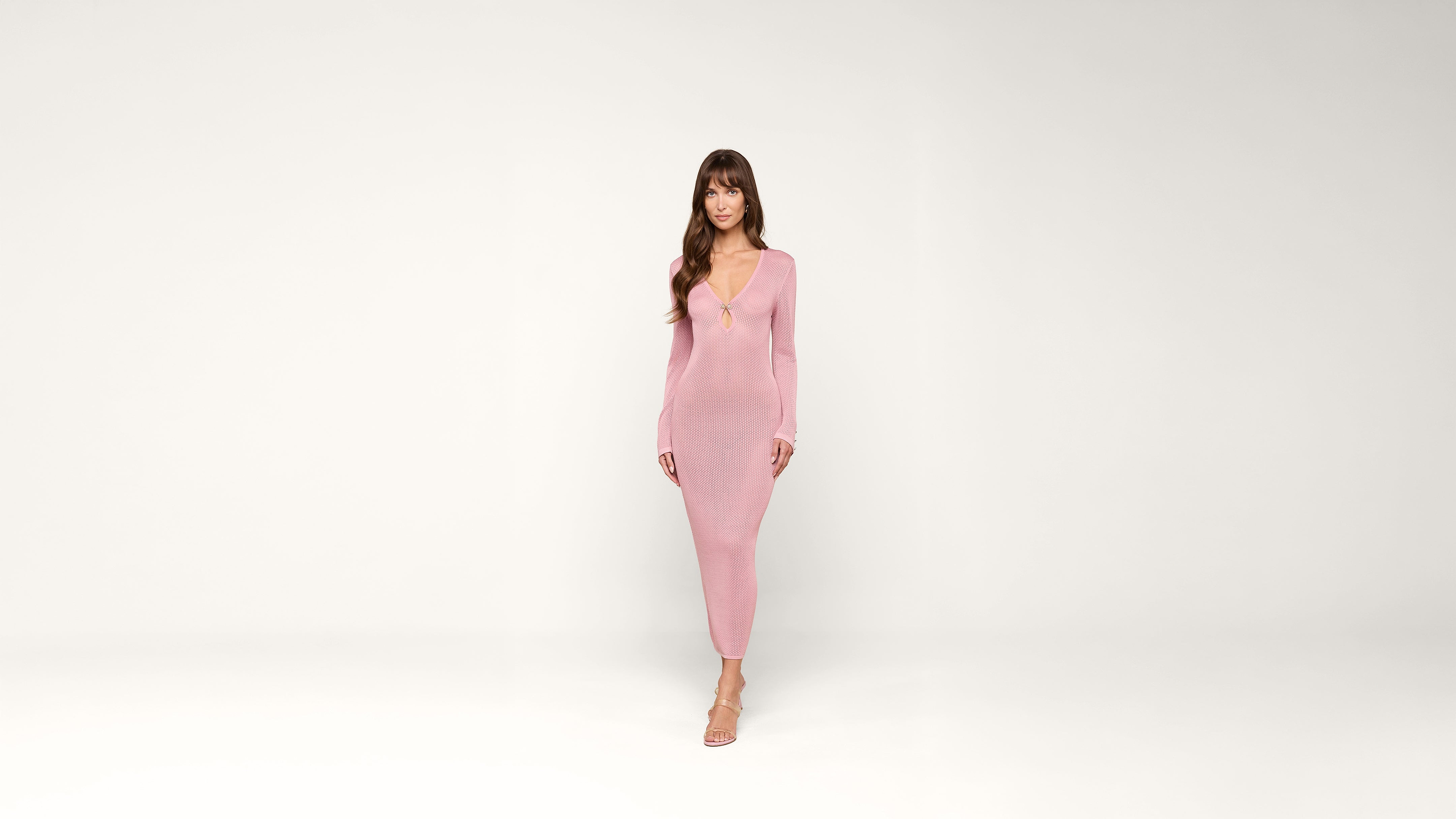 A woman in a long-sleeved, form-fitting pink dress and heels stands against a plain white background, facing forward with a neutral expression.