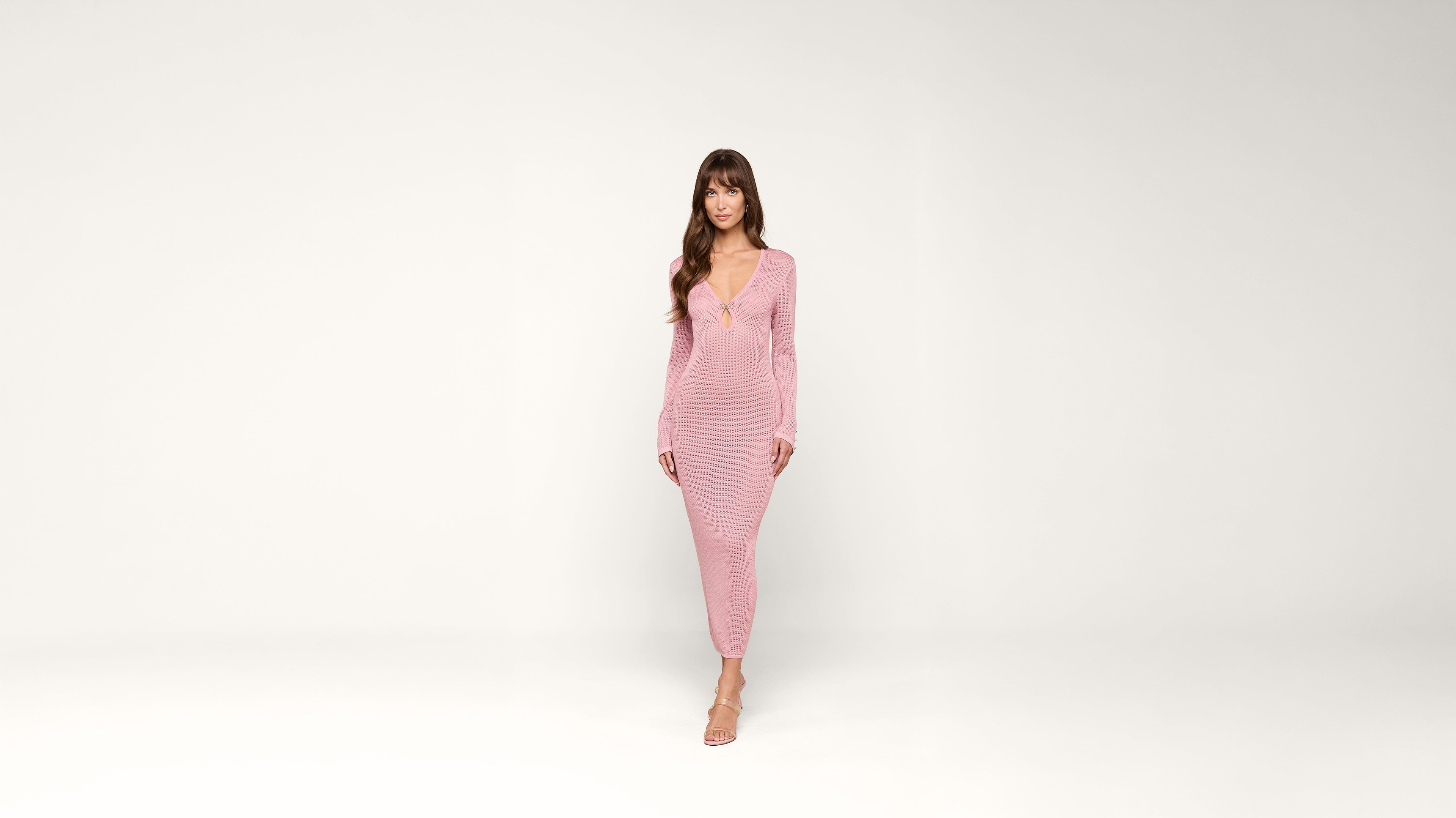 A woman in a long-sleeved, form-fitting pink dress and heels stands against a plain white background, facing forward with a neutral expression.