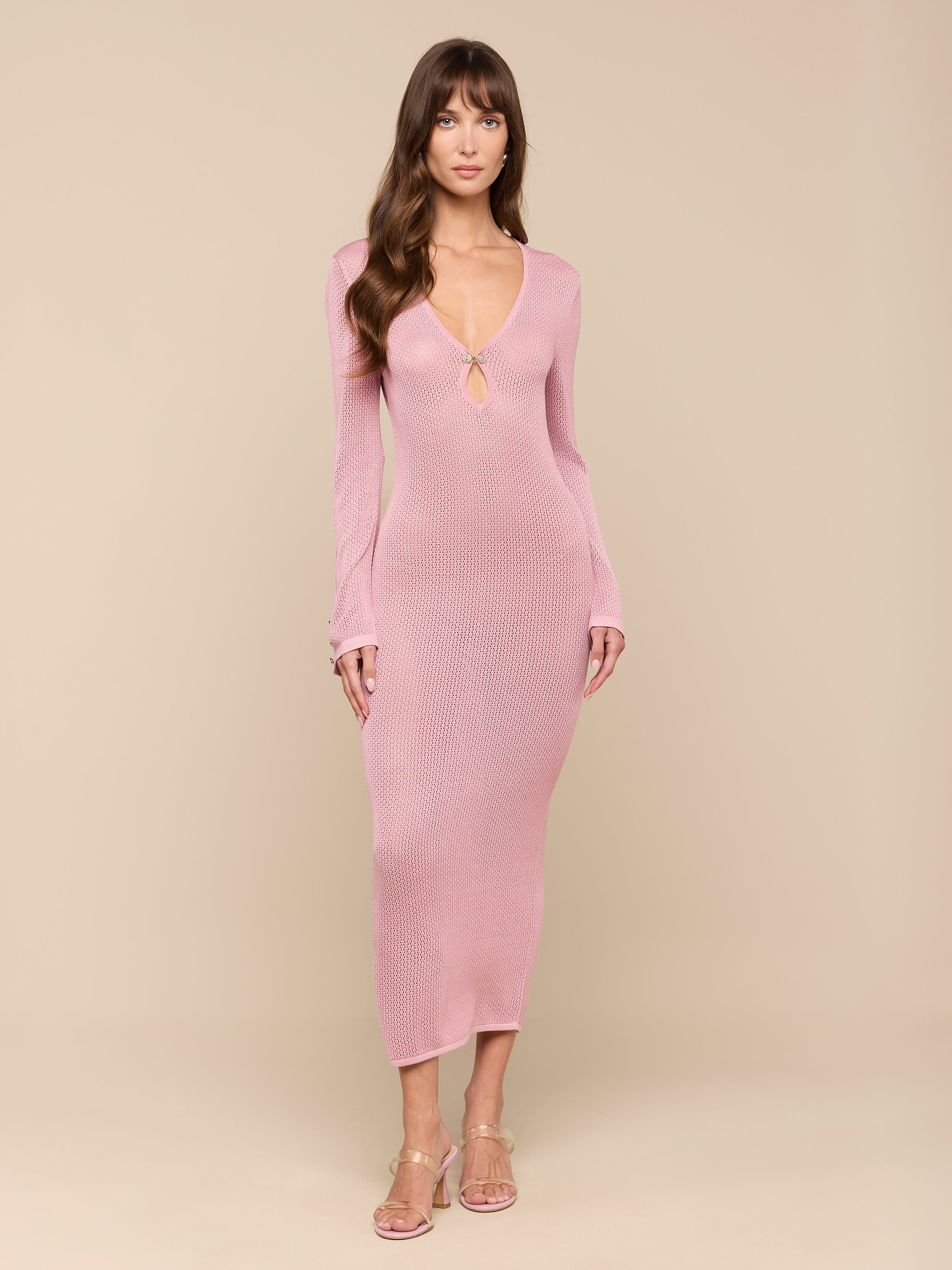 A woman wearing the Sydney Knit Dress—a long-sleeve, light pink, form-fitting maxi—stands against a plain white background, looking forward. She pairs the dress with clear heeled sandals and has long brown hair with bangs.