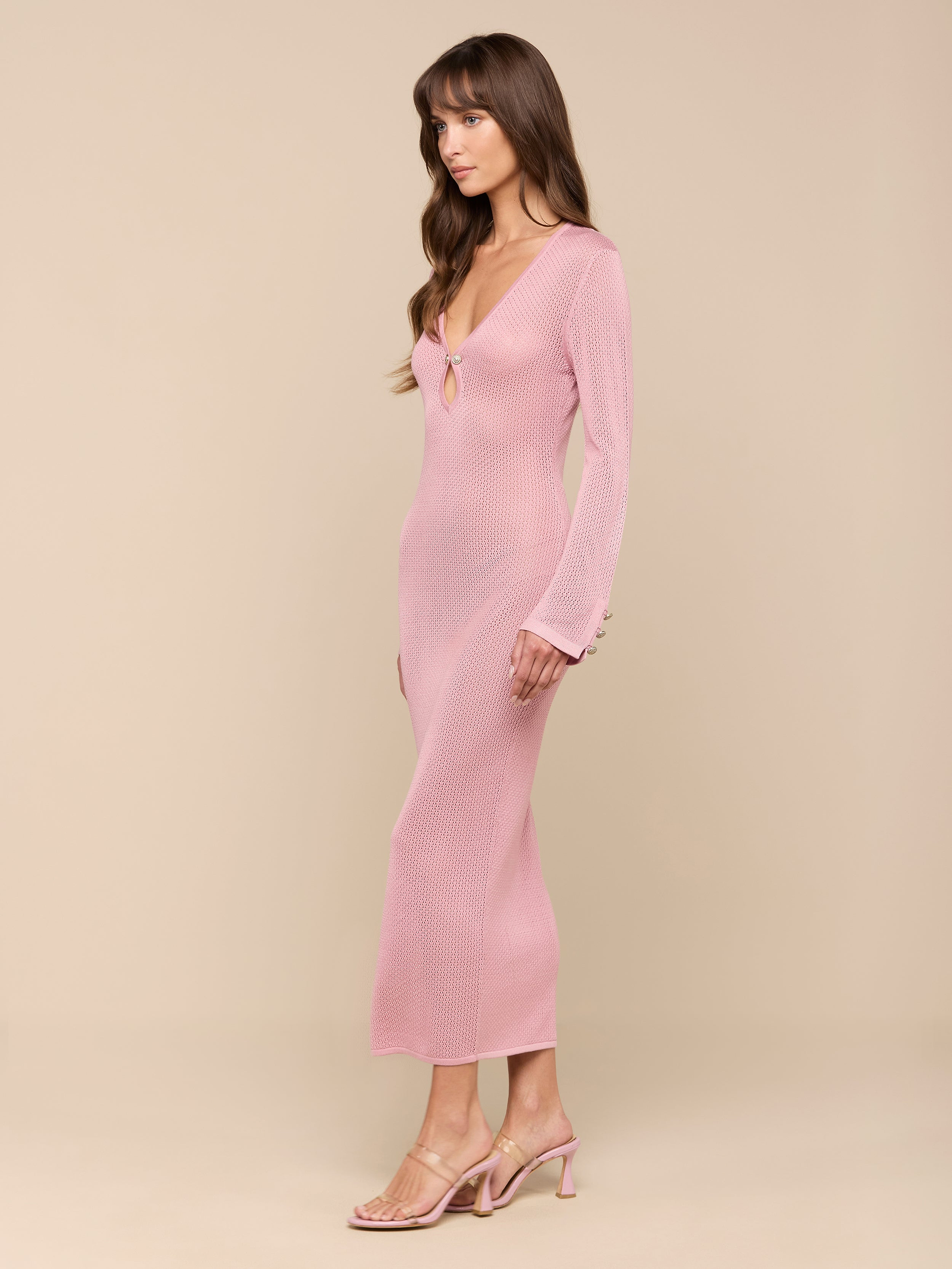A woman in a fitted, long-sleeve pink Sydney Knit Dress and heeled sandals stands against a plain white background, posed in profile with her arms relaxed by her sides.