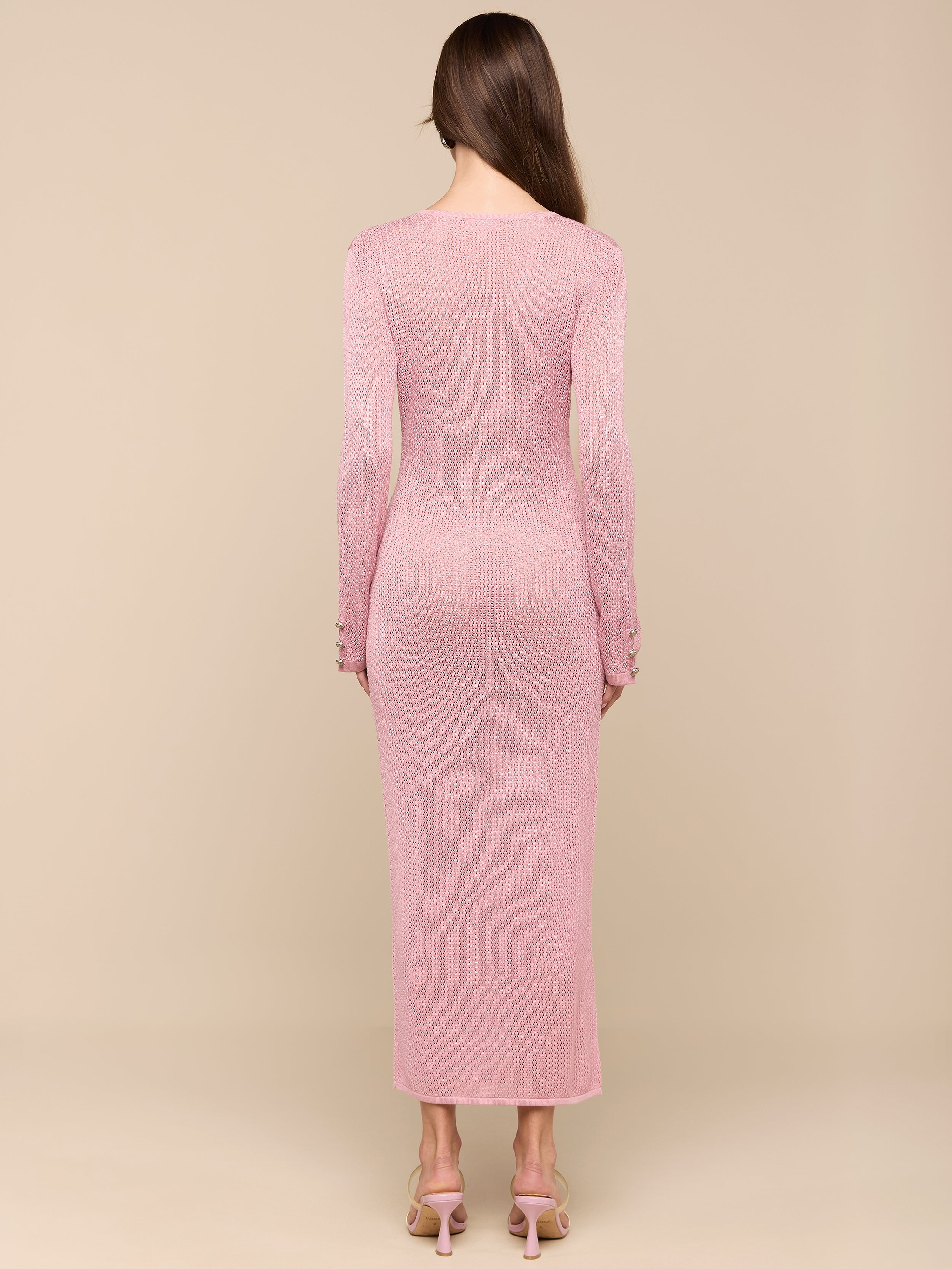 A woman stands facing away, wearing a long-sleeved, ankle-length Sydney Knit Dress in pink with heeled sandals. She has straight brown hair and stands against a plain white background.