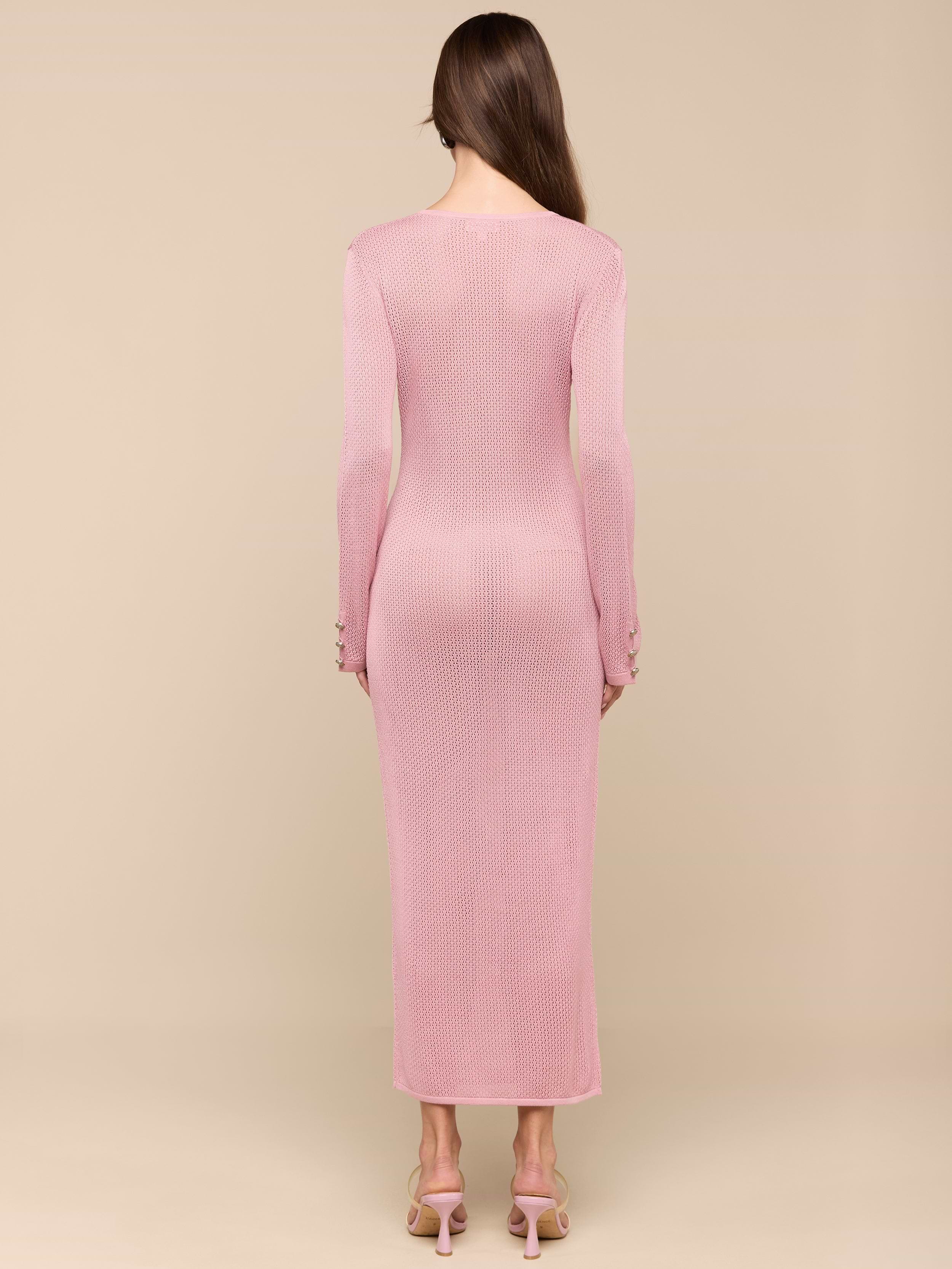 A woman stands facing away, wearing a long-sleeved, ankle-length Sydney Knit Dress in pink with heeled sandals. She has straight brown hair and stands against a plain white background.