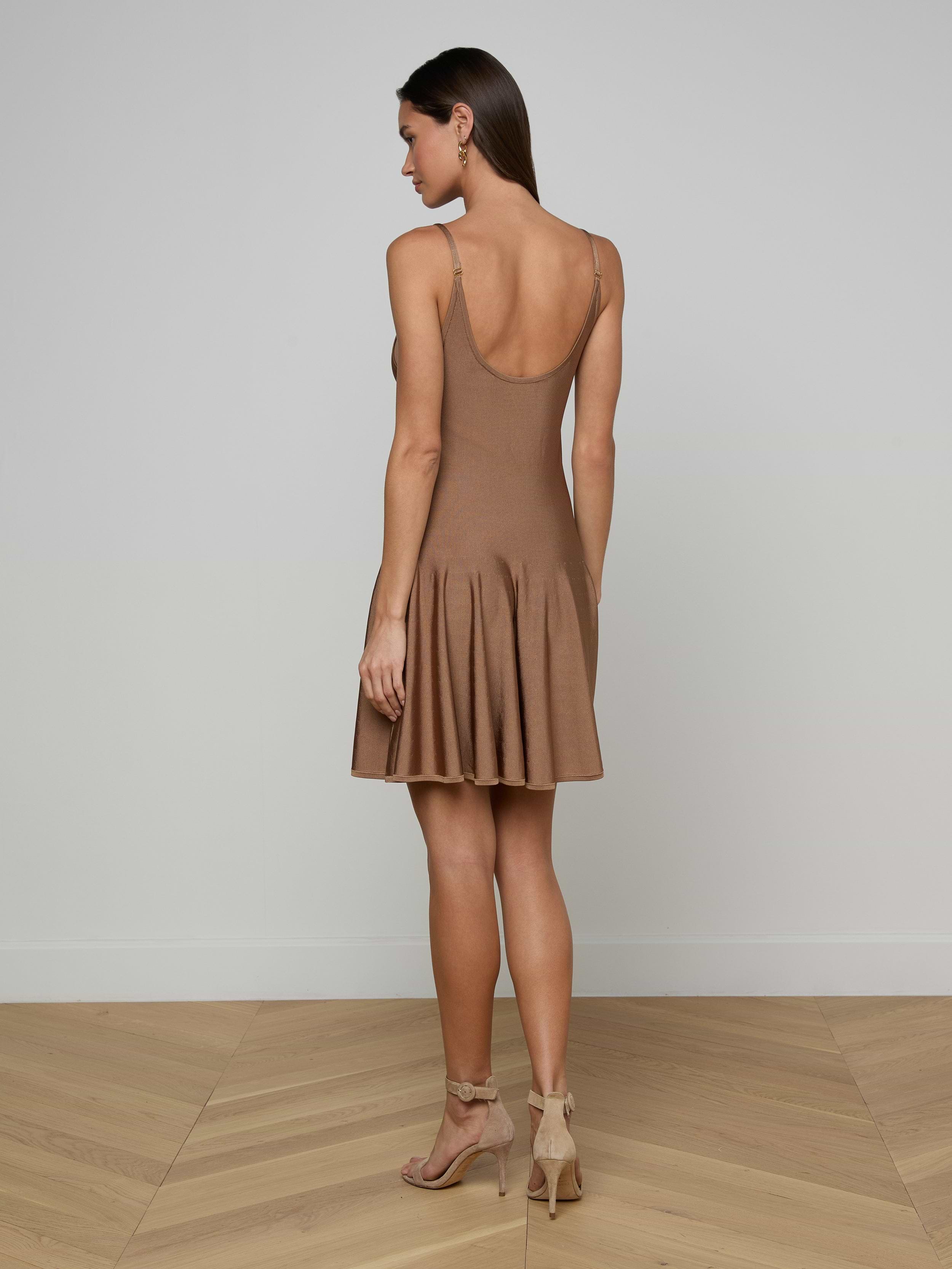 A woman stands facing away against a plain white background, wearing the Lana Knit Dress—a sleeveless brown stretch knit design with a flared skirt—paired with beige high-heeled sandals.