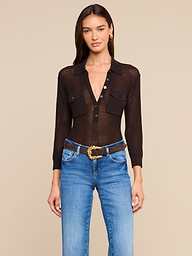 A woman wears the Carine Top, a sheer black button-up shirt with front pockets, paired with blue jeans and a decorative belt, styled for Fall 2026 and posed against a plain light background.