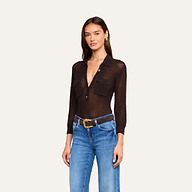 A woman wearing the Carine Top—a lightweight knit sheer black long-sleeve button-up with front pockets—paired with blue jeans, a brown belt with a gold buckle, and gold hoop earrings, stands against a plain light background.