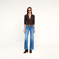 A woman wearing sunglasses, a sheer black button-up top layered over the lightweight knit Carine Top, blue jeans with a brown belt, and brown heeled boots stands against a plain white background.