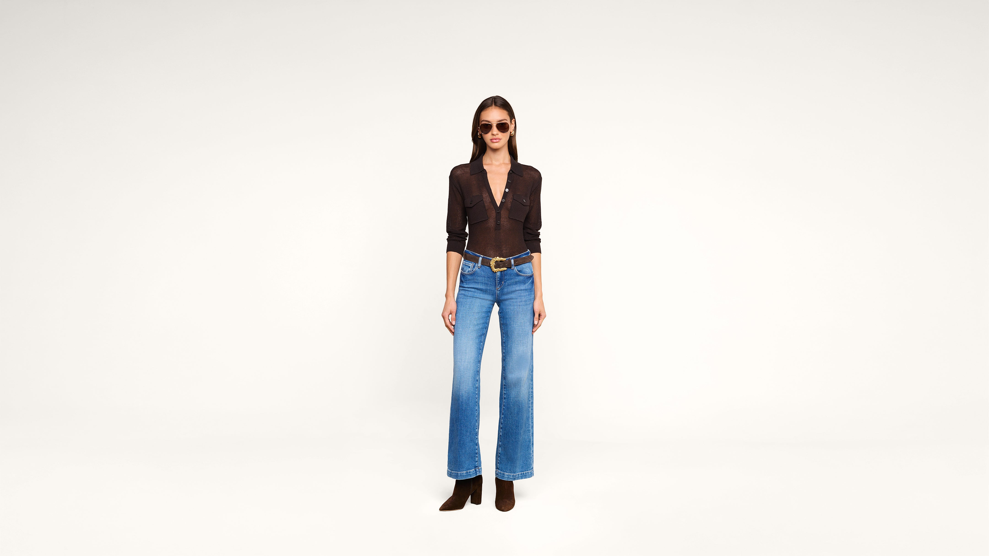 Woman wearing sunglasses, a dark button-down shirt, blue high-waisted wide-leg jeans, a brown belt, and brown boots, standing against a plain white background.
