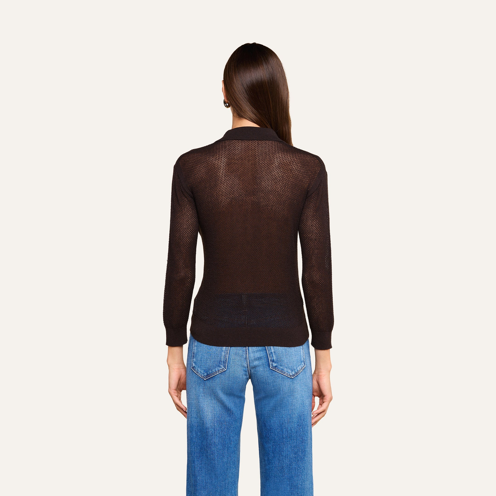 A person stands with their back to the camera, wearing a sheer black Carine Top and blue jeans against a plain light background, capturing a sleek Fall 2026 vibe.