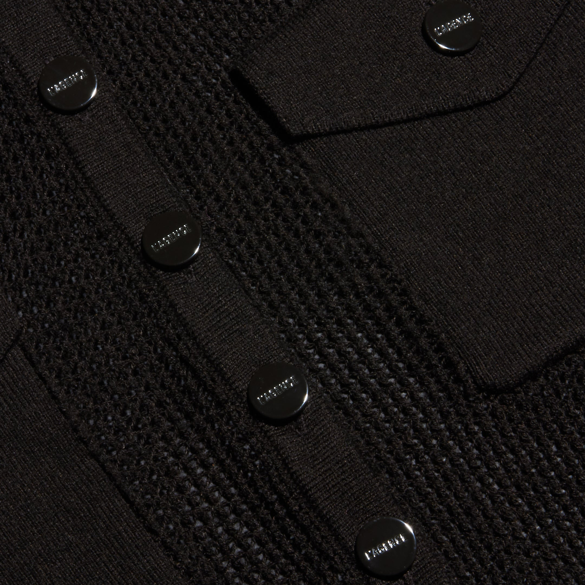Close-up of a black lightweight knit Carine Top featuring shiny round buttons engraved with the word PRADA and a flap pocket. Perfect for Fall 2026.