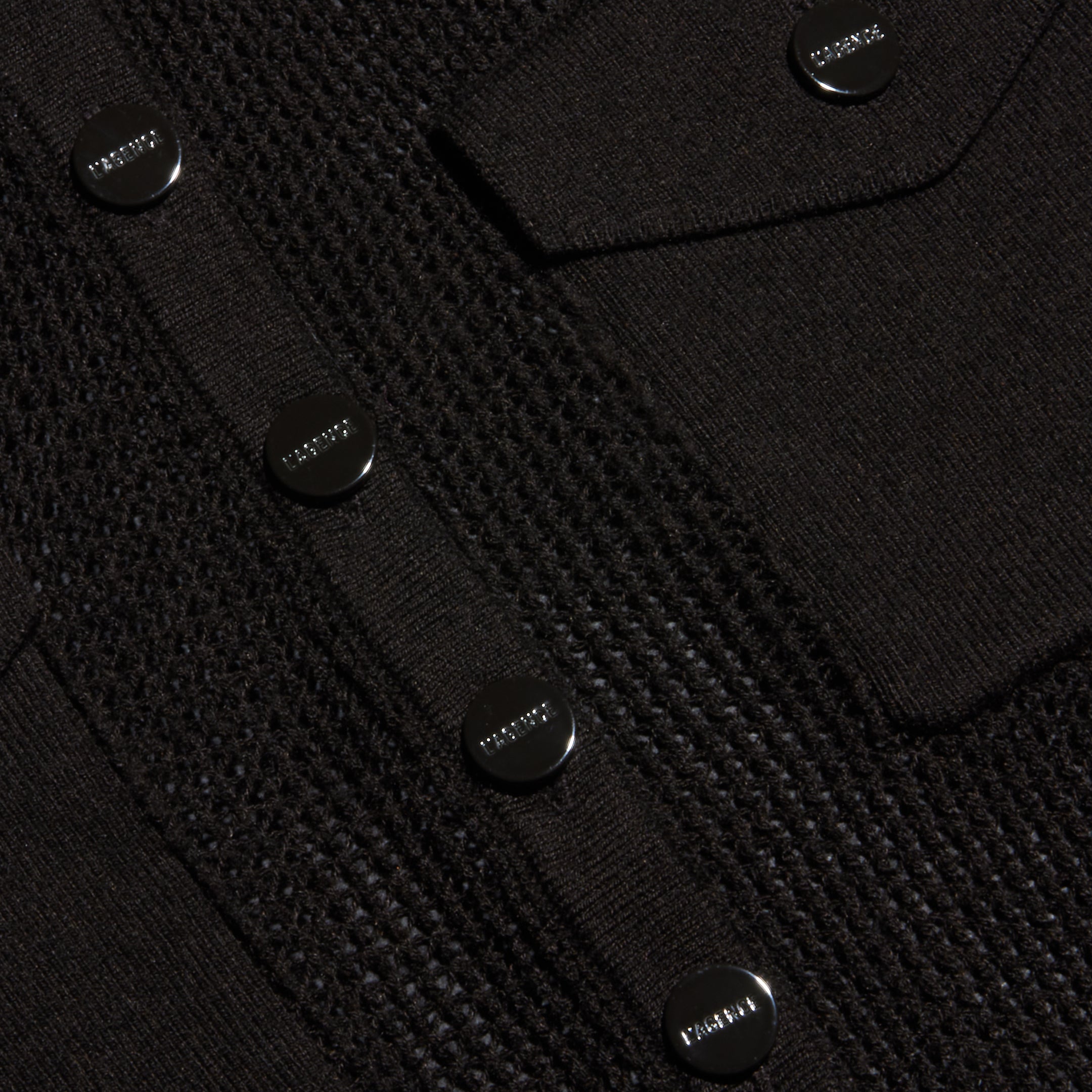 Close-up of a black lightweight knit Carine Top featuring shiny round buttons engraved with the word PRADA and a flap pocket. Perfect for Fall 2026.
