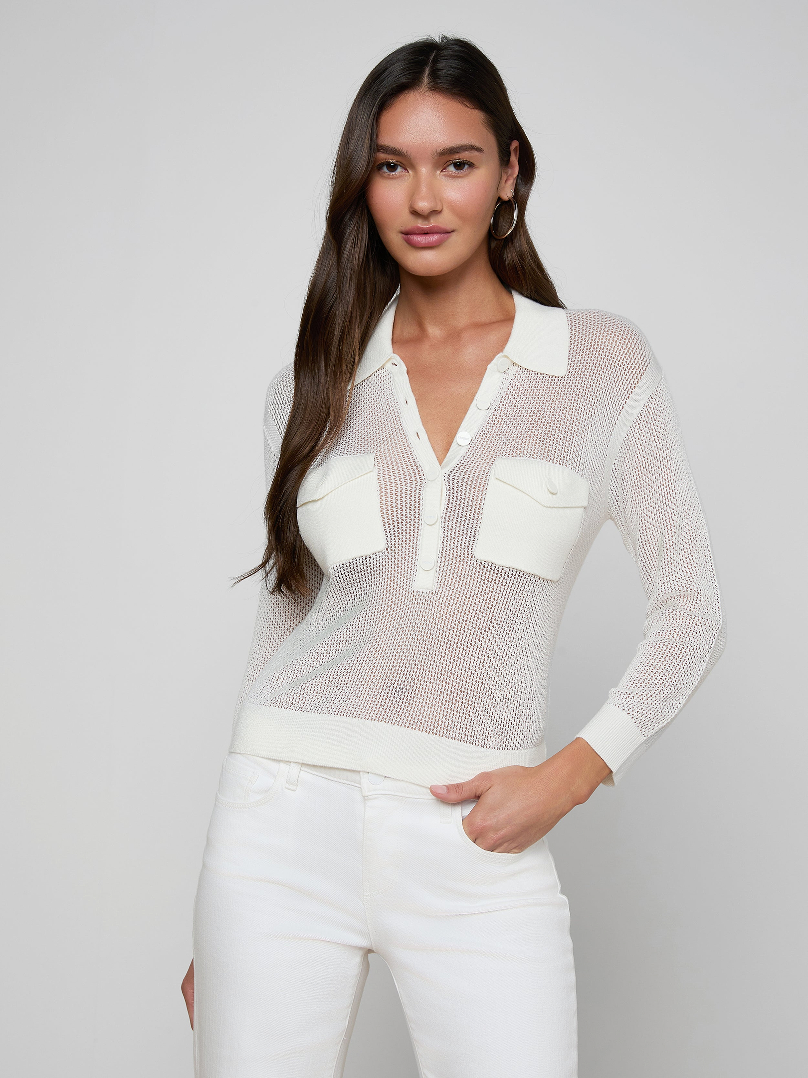 A woman with long dark hair wears the Carine Top—a sheer, white, long-sleeve collared shirt made from lightweight knit fabric with utility pockets—paired with white pants, standing against a plain light background with one hand in her pocket.