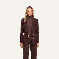 A woman stands against a plain, light background wearing the Amelia Knit Blazer in dark brown with gold buttons, paired with a matching brown turtleneck and trousers. Her long blonde hair and neutral expression highlight the soft tailoring of the look.