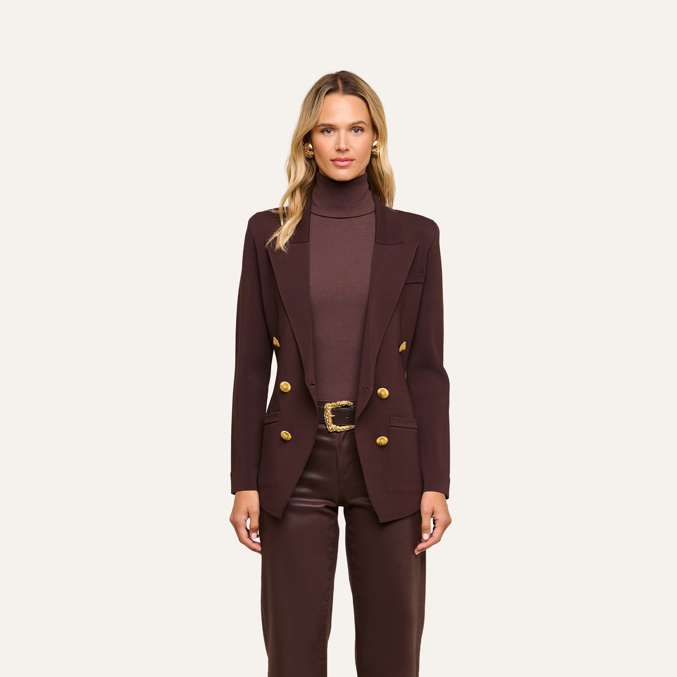 A woman stands against a plain, light background wearing the Amelia Knit Blazer in dark brown with gold buttons, paired with a matching brown turtleneck and trousers. Her long blonde hair and neutral expression highlight the soft tailoring of the look.