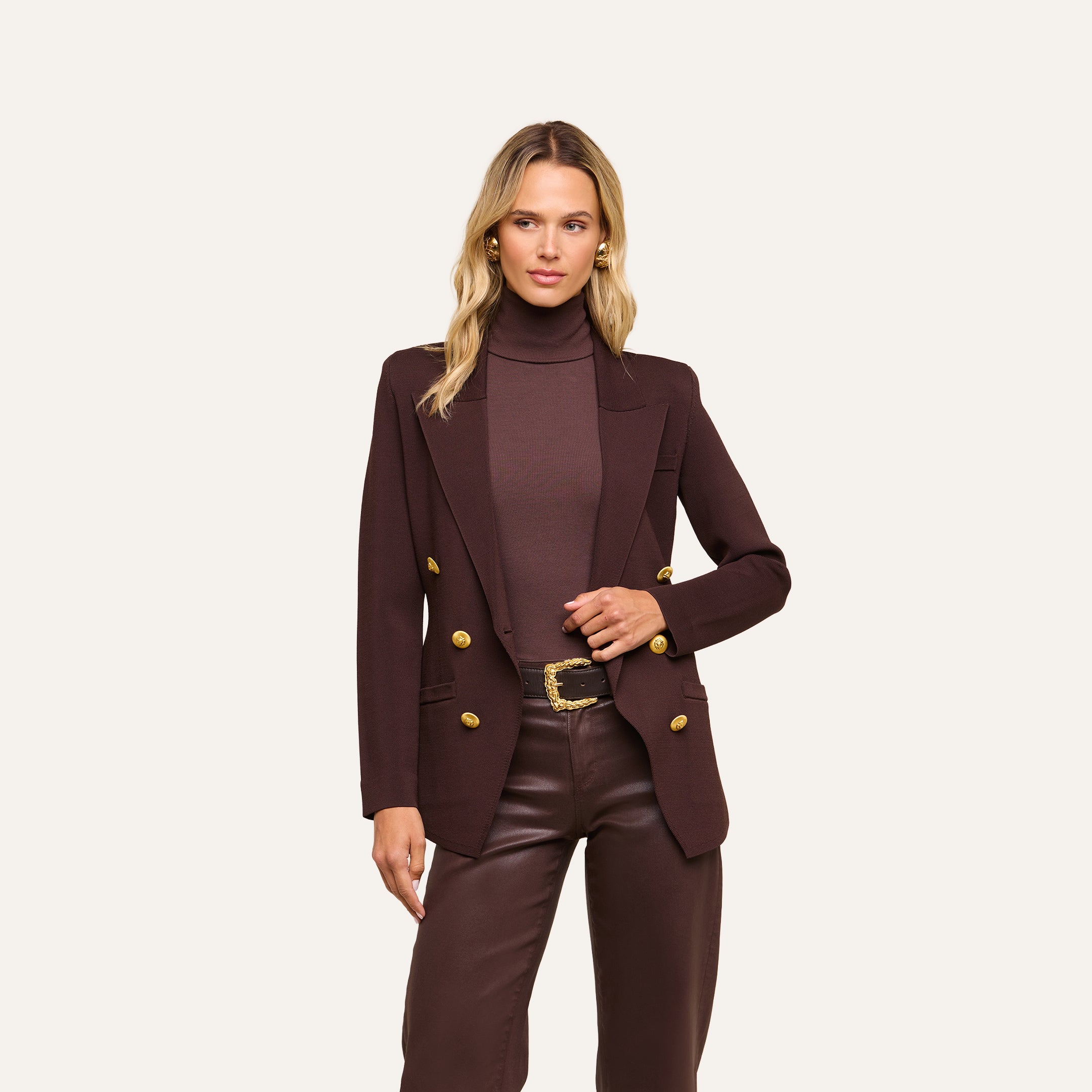 Wearing the Amelia Knit Blazer in dark brown with gold buttons and soft tailoring, paired with a matching brown turtleneck and brown leather pants, a woman stands against a plain light background.