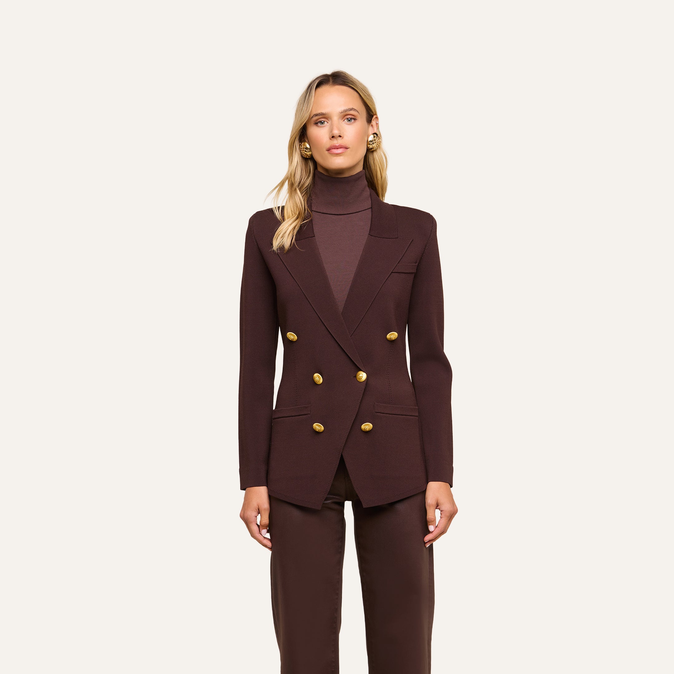 Wearing the Amelia Knit Blazer, a woman stands facing forward against a plain, light background, styled with gold buttons and paired with matching brown pants.