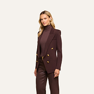 A woman with blonde hair wears the Amelia Knit Blazer—a dark brown structured knit piece with gold buttons—paired with a matching turtleneck and brown pants, standing against a plain light background.