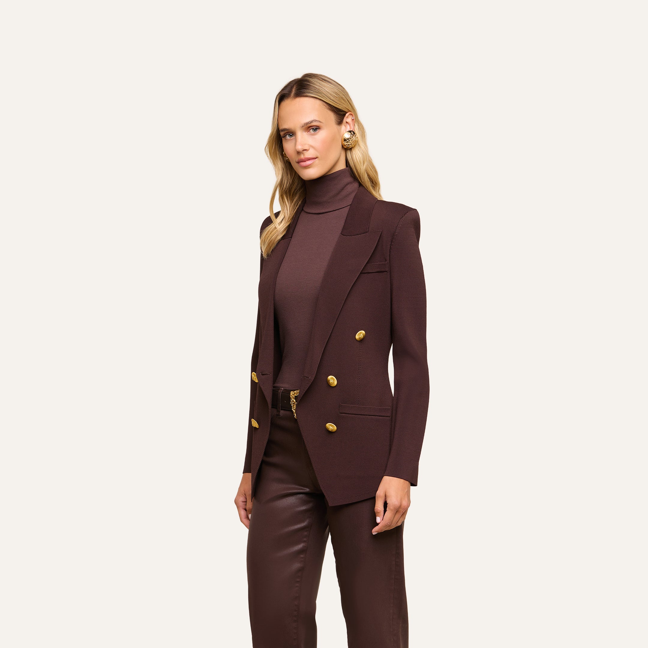A woman with blonde hair wears the Amelia Knit Blazer—a dark brown structured knit piece with gold buttons—paired with a matching turtleneck and brown pants, standing against a plain light background.