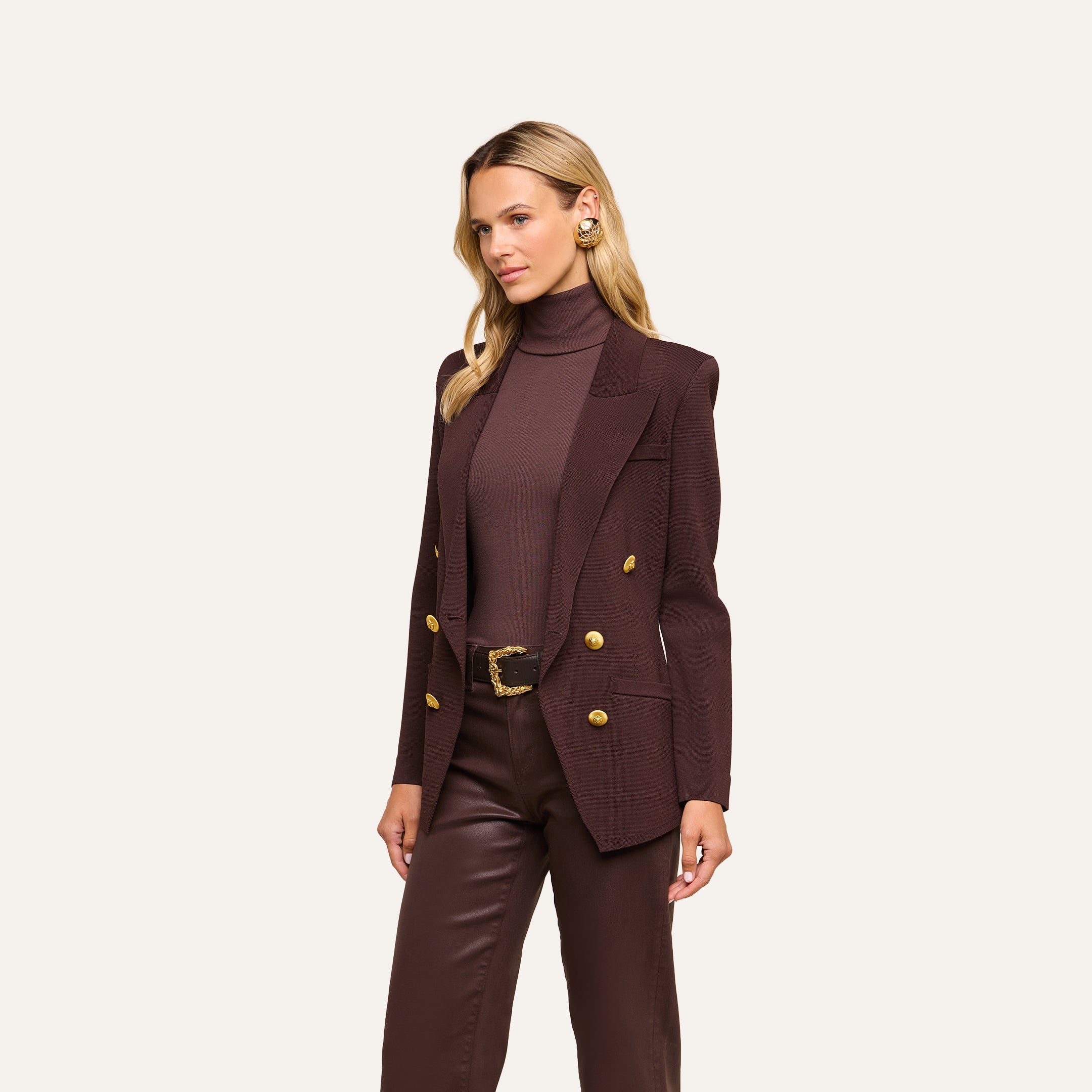 A woman with blonde hair wears the Amelia Knit Blazer in dark brown, featuring gold buttons, paired with a matching turtleneck and brown leather pants. She stands against a plain light background, facing slightly to the side.