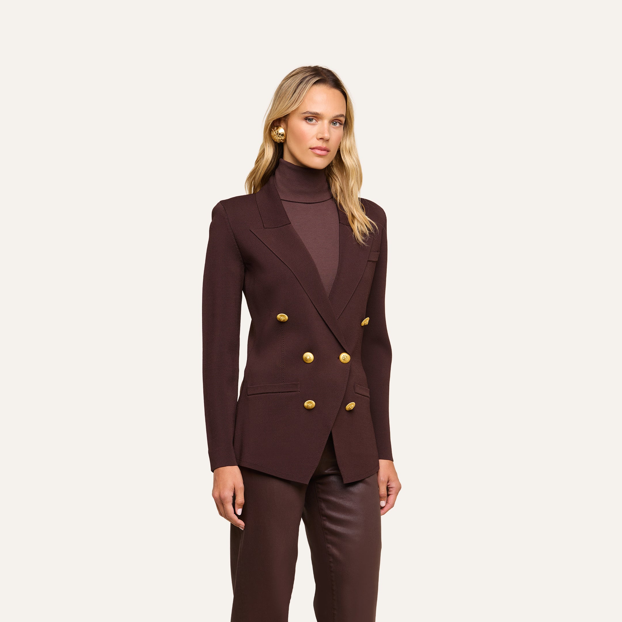 Wearing the Amelia Knit Blazer in dark brown with gold buttons, a woman stands against a plain light background, layered over a matching brown turtleneck and pants.