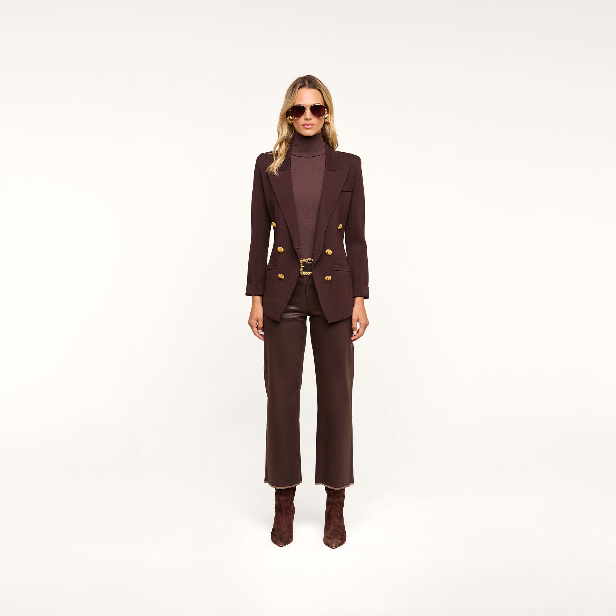 A woman stands facing forward against a plain light background, wearing sunglasses, brown pants, brown heeled boots, a brown turtleneck, and the Amelia Knit Blazer with gold buttons.